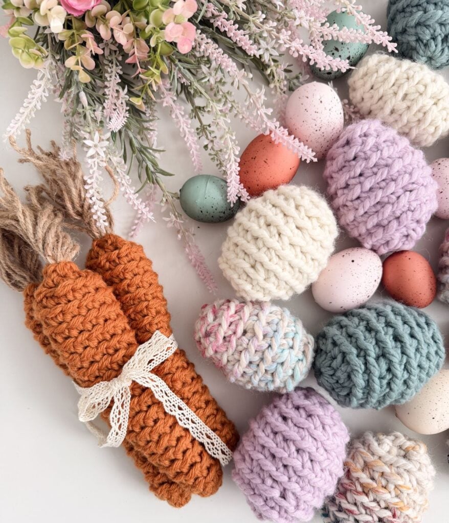A collection of crocheted Easter eggs in various colors, three crocheted carrots tied with lace, and artificial flowers, all arranged on a white surface.