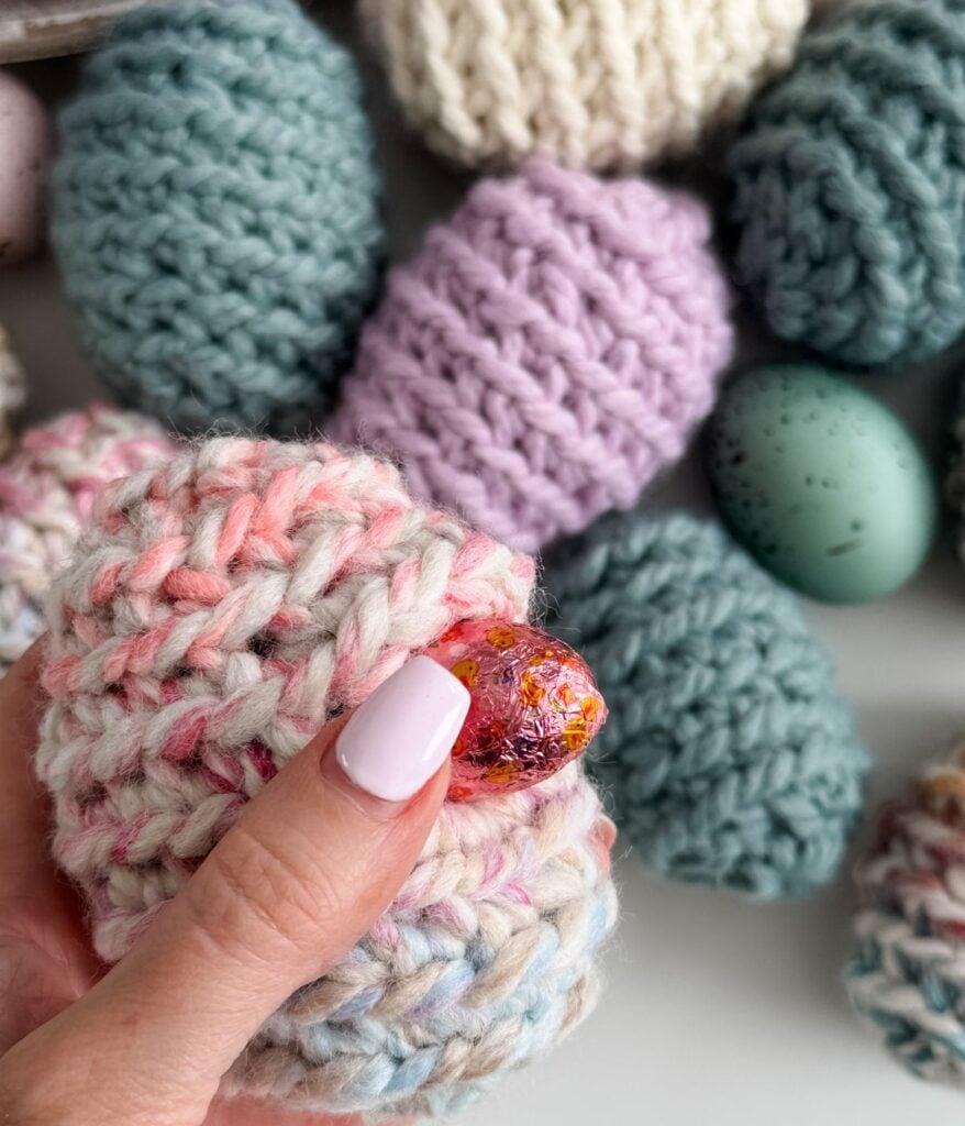A hand with light pink nail polish holds a small, pink-wrapped Easter Egg inside a knitted pastel egg-shaped pouch, surrounded by other knitted pouches and a speckled egg.