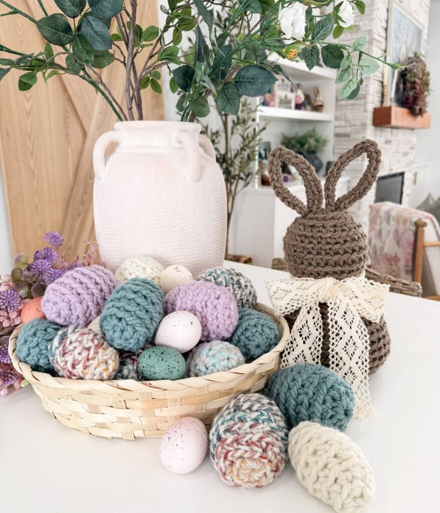 A basket of colorful knitted and speckled Easter eggs sits on a white table next to a crocheted bunny with a lace bow, a white vase, and green foliage.