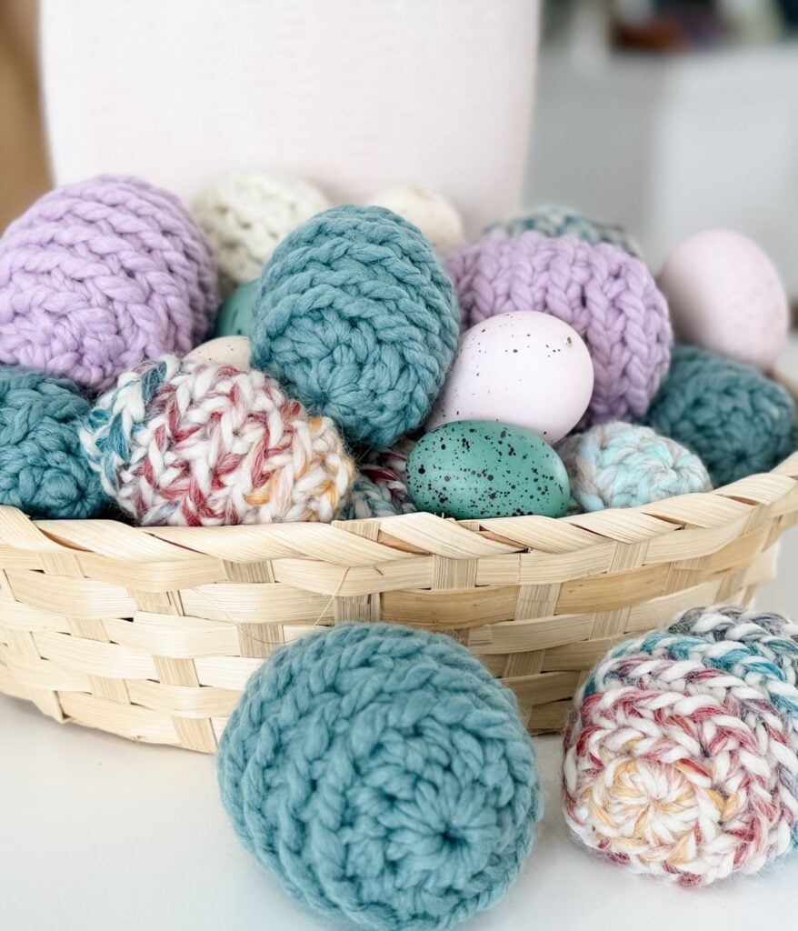 A woven basket filled with colorful knitted and painted Easter eggs, with a few eggs placed on the table nearby.