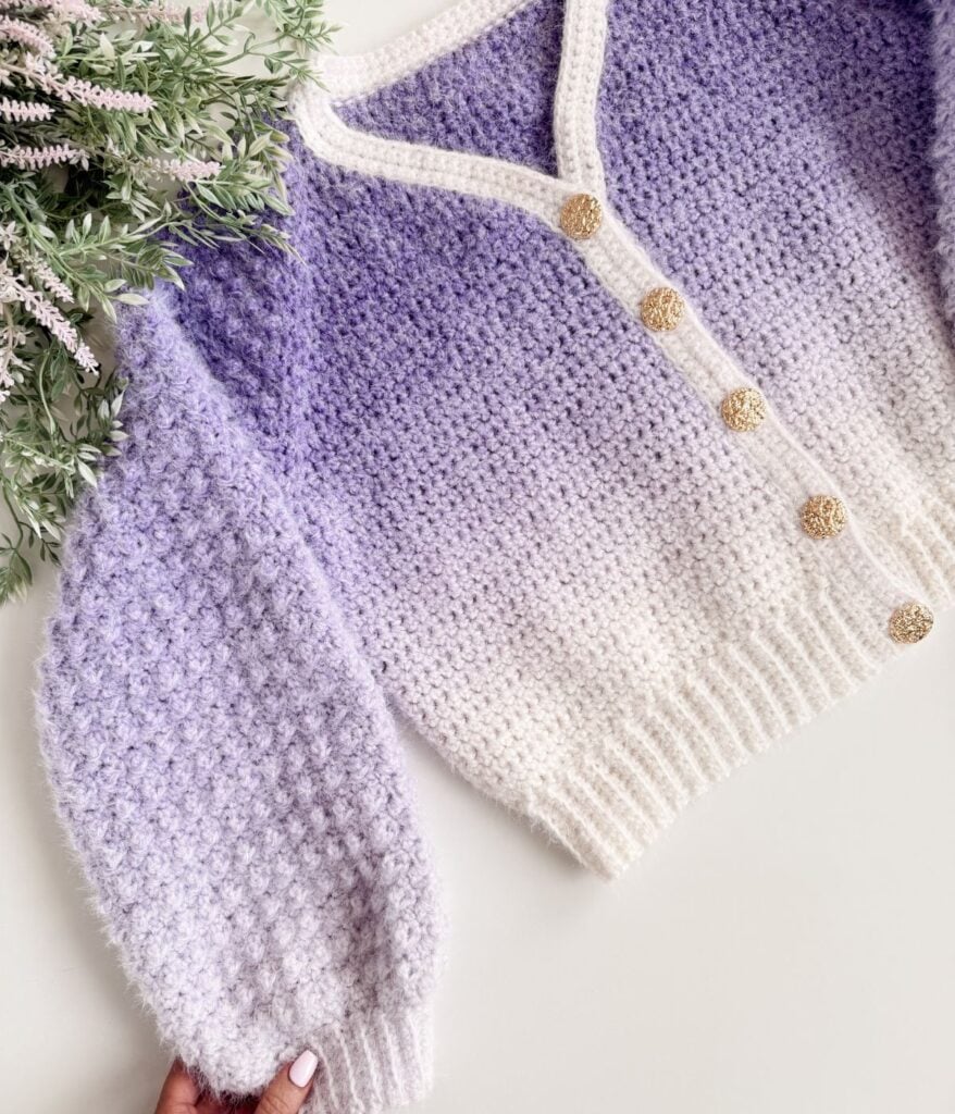 A knitted cardigan with a gradient pattern from white to lavender, gold buttons, and a textured stitch, displayed beside green foliage—perfect for those seeking a unique Cardigan Crochet Pattern.