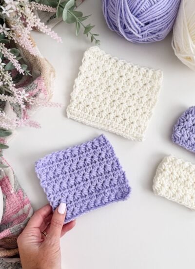 A hand holds a lavender crocheted swatch next to other white and lavender swatches, yarn balls, and flowers on a white surface—perfect for anyone wanting to Learn Star Stitch or follow a Crochet Star Stitch tutorial.