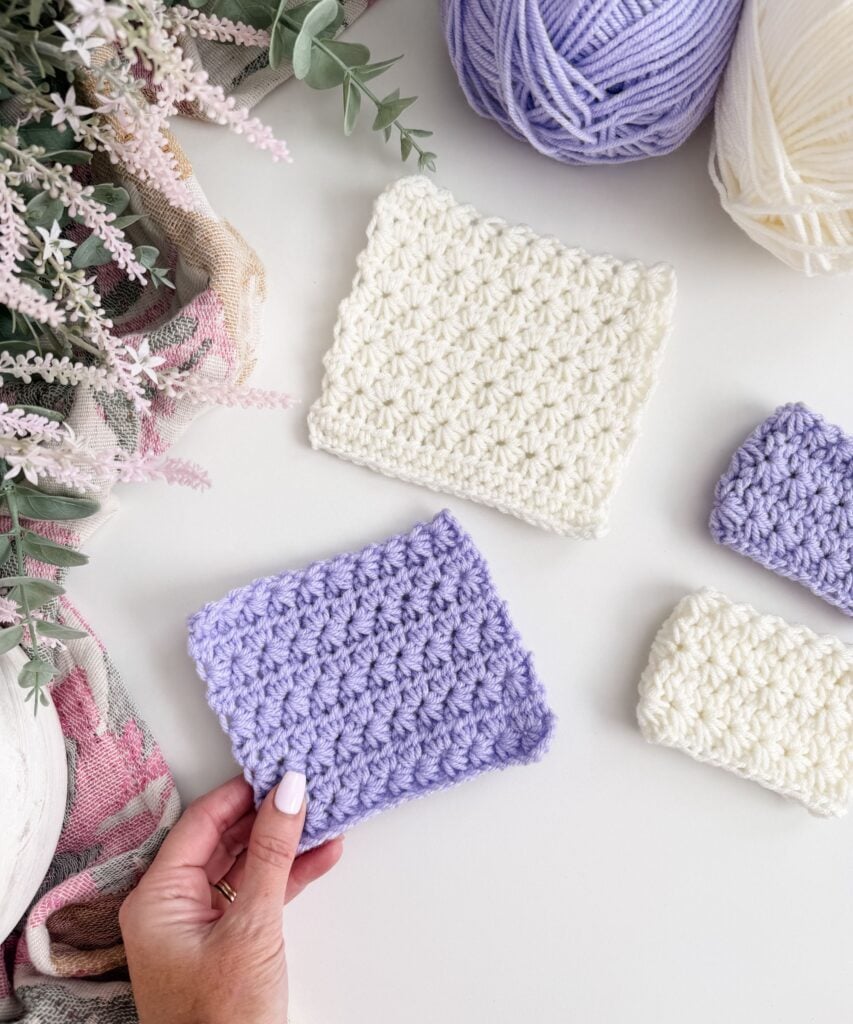 A hand holds a lavender crocheted swatch next to other white and lavender swatches, yarn balls, and flowers on a white surface—perfect for anyone wanting to Learn Star Stitch or follow a Crochet Star Stitch tutorial.