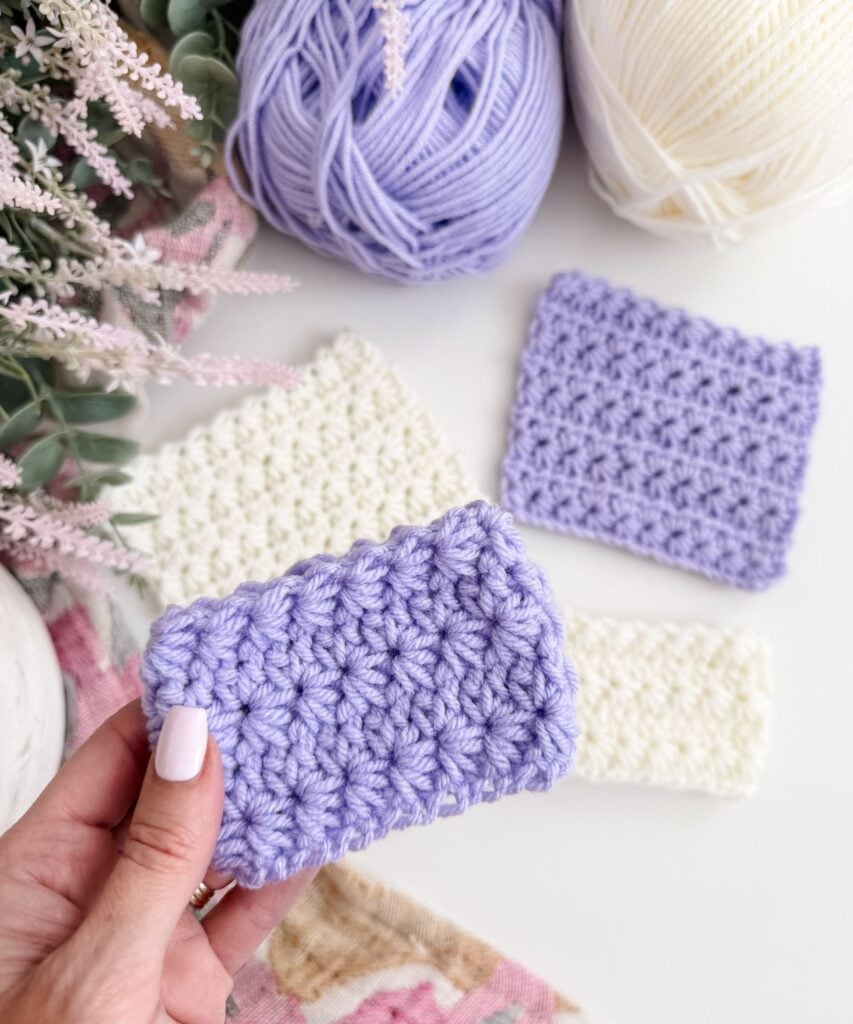 A hand holds a small swatch of lavender Crochet Star Stitch. Two similar white swatches and one lavender swatch are laid on a surface next to balls of yarn and some decorative greenery.