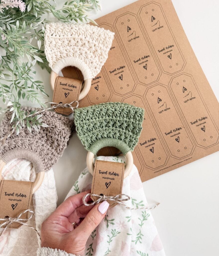 Hand holding a crocheted baby teether with wooden ring, two more in beige and green nearby—perfect for gifting alongside handmade towel holder tags—on a white surface with greenery.