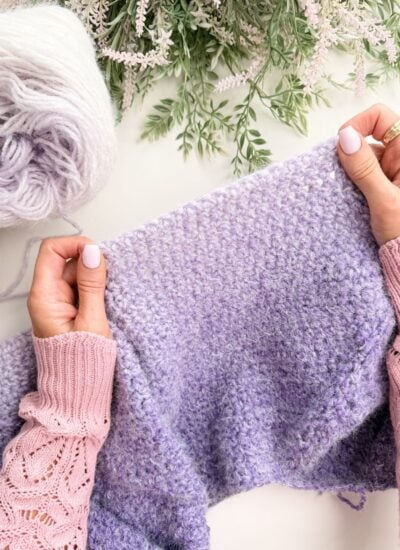 Hands holding knitting needles, working on a lavender-colored textured knit piece, with a ball of matching yarn and greenery in the background—perfect for practicing crochet abbreviations and symbols.