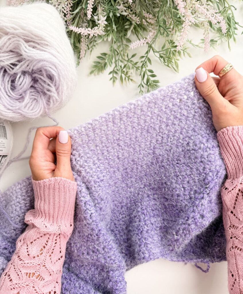 Hands holding knitting needles, working on a lavender-colored textured knit piece, with a ball of matching yarn and greenery in the background—perfect for practicing crochet abbreviations and symbols.