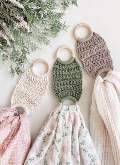 Three crocheted teething rings with attached patterned muslin cloths, which can also double as a soft towel holder, are laid out on a white surface beside green leafy sprigs.