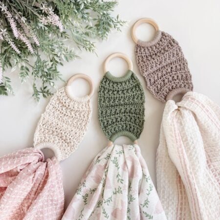Three crocheted teething rings with attached patterned muslin cloths, which can also double as a soft towel holder, are laid out on a white surface beside green leafy sprigs.