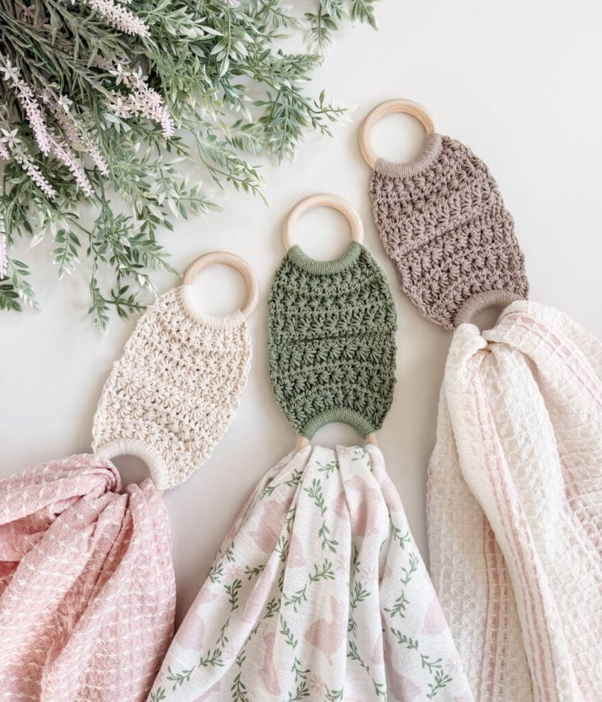 Three crocheted teething rings with attached patterned muslin cloths, which can also double as a soft towel holder, are laid out on a white surface beside green leafy sprigs.