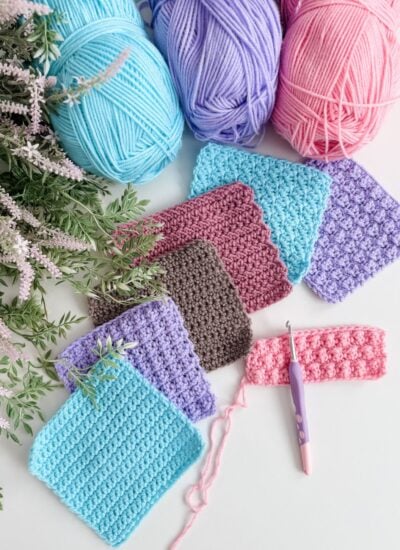 Several crochet swatches in different colors and textures are laid out beside yarn balls in blue, purple, and pink, along with a crochet hook, artificial lavender, and a guide to crochet abbreviations and symbols on a white surface.