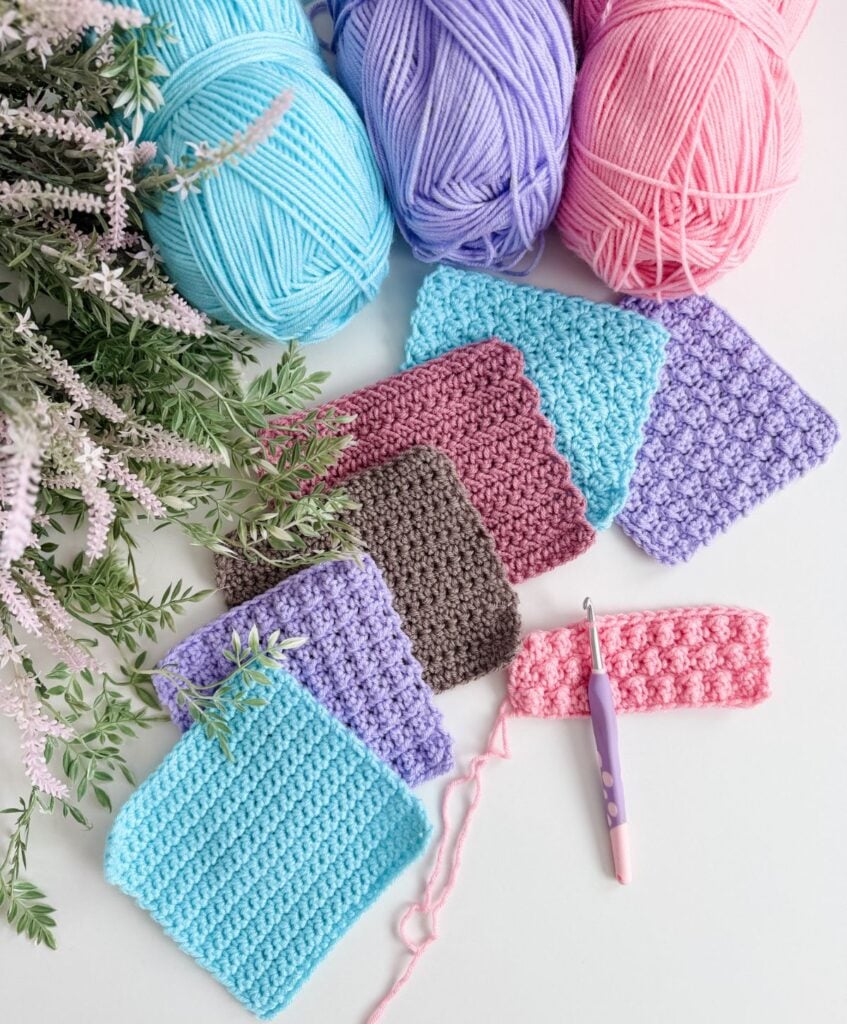 Several crochet swatches in different colors and textures are laid out beside yarn balls in blue, purple, and pink, along with a crochet hook, artificial lavender, and a guide to crochet abbreviations and symbols on a white surface.