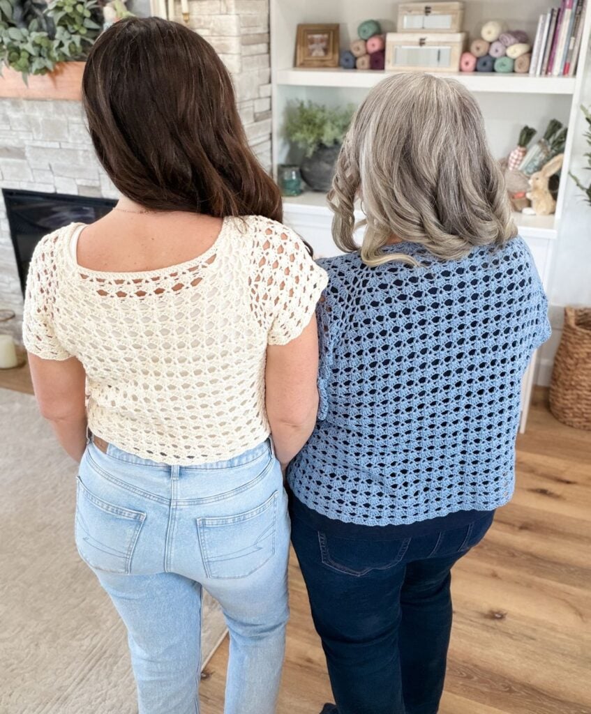 Two women stand indoors with their backs to the camera, modeling crocheted tops—one in cream and one in blue—crafted with a beautiful shell stitch, in front of shelves filled with yarn and books.