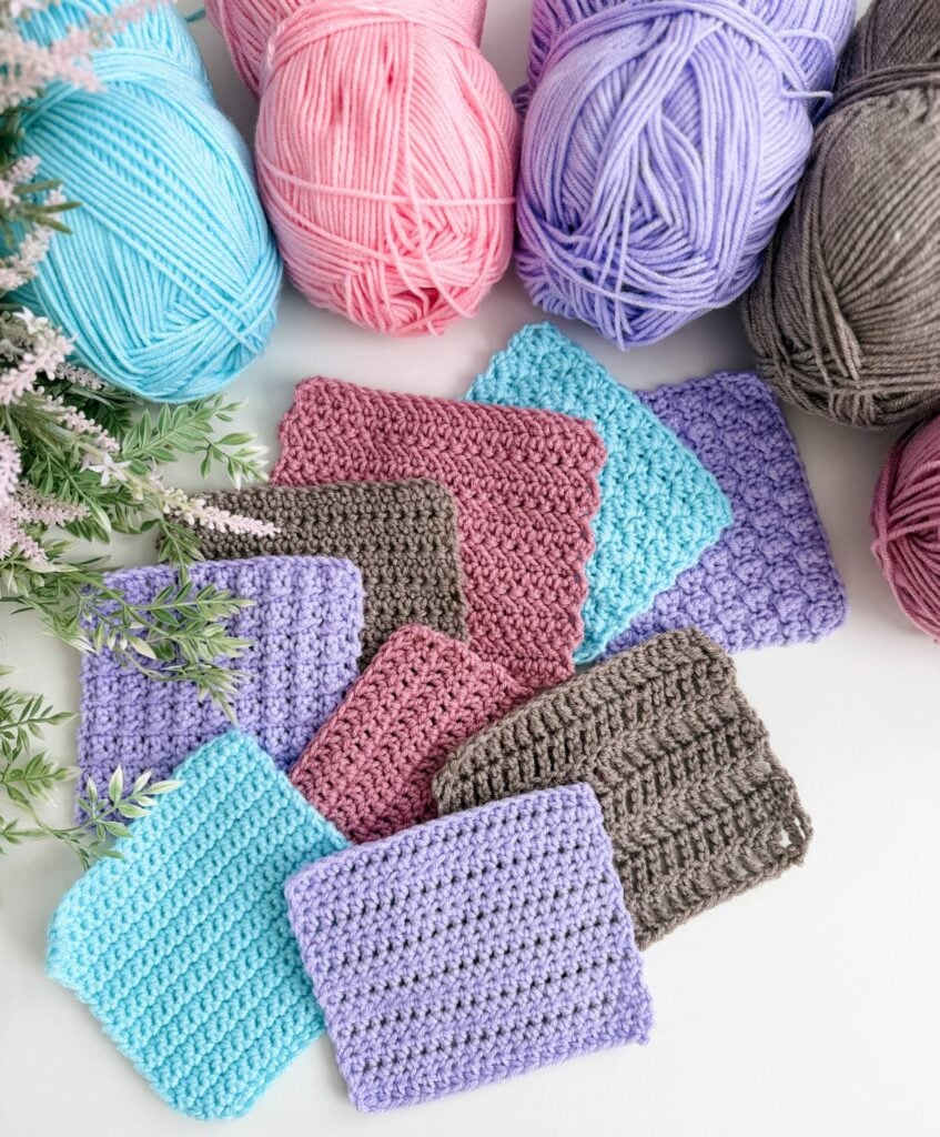 Several crocheted swatches in pastel colors are arranged on a white surface, surrounded by skeins of pink, purple, blue, and brown yarn, with some greenery on the side—perfect for practicing crochet abbreviations and symbols.