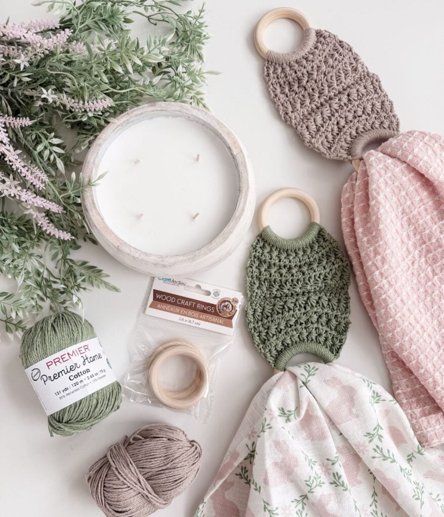 Yarn, wood craft rings, a white candle, a towel holder, artificial greenery, and two textured baby loveys with wooden rings are arranged on a light surface.