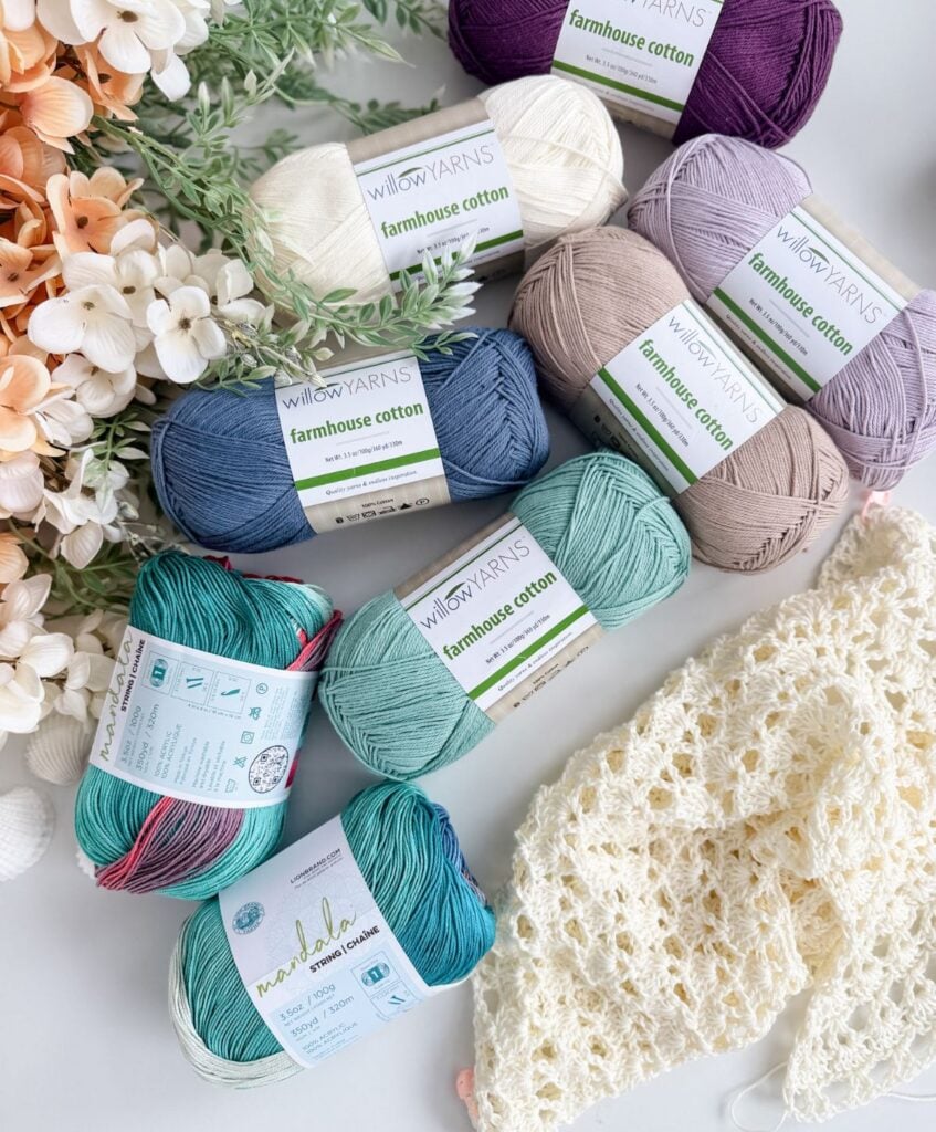 Several skeins of cotton yarn in various colors are arranged with artificial flowers and a partially crocheted cream piece featuring a beautiful Shell Stitch Crochet, perfect for making your next Solea crochet cardigan pattern.