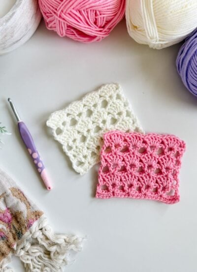Two crocheted swatches, one white and one pink, are displayed on a white surface with balls of yarn, a crochet hook, and patterned fabric nearby—perfect for following a shells crochet or V-stitch crochet tutorial.