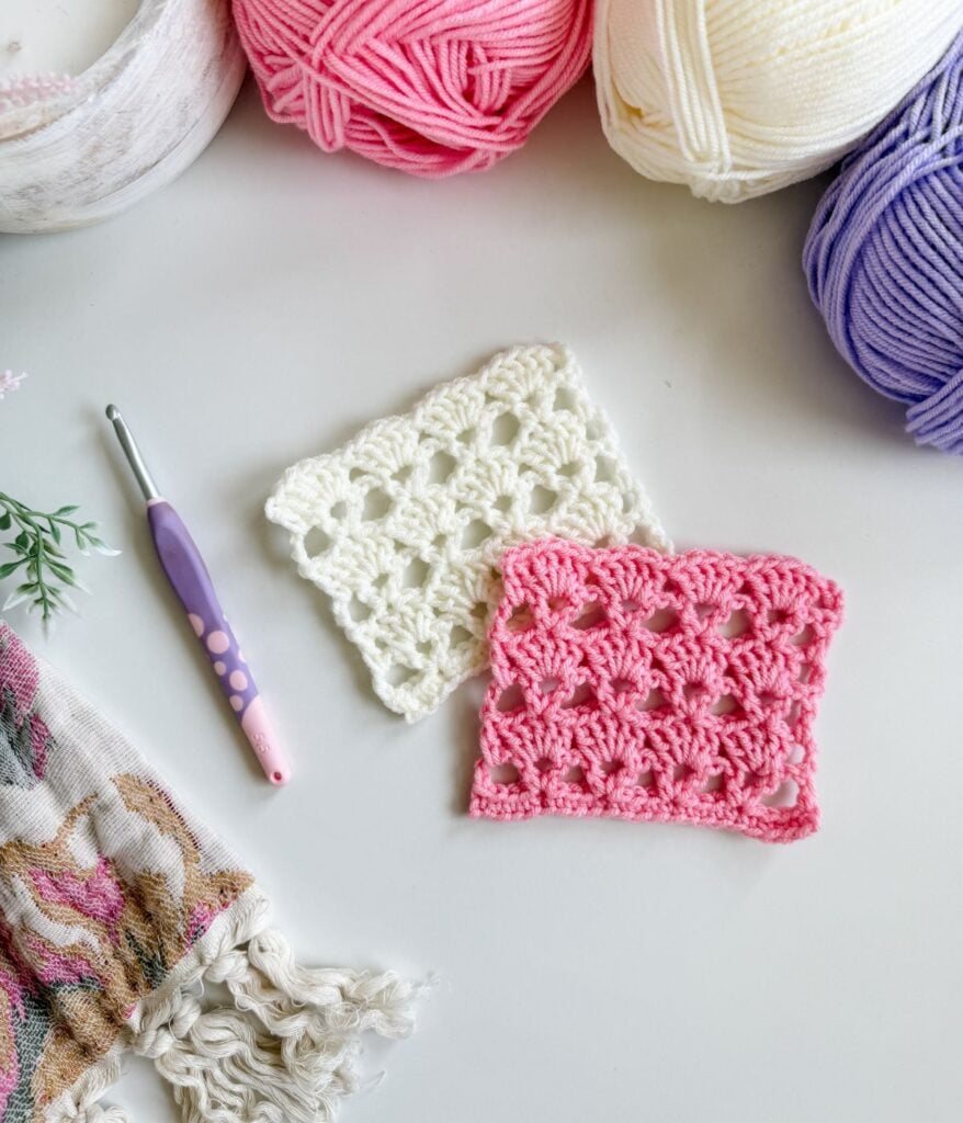 Two crocheted swatches, one white and one pink, are displayed on a white surface with balls of yarn, a crochet hook, and patterned fabric nearby—perfect for following a shells crochet or V-stitch crochet tutorial.