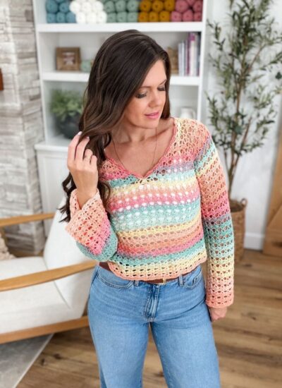 Woman wearing a colorful, shell stitch crocheted sweater and blue jeans stands indoors, touching her hair, with shelves of yarn and plants in the background.