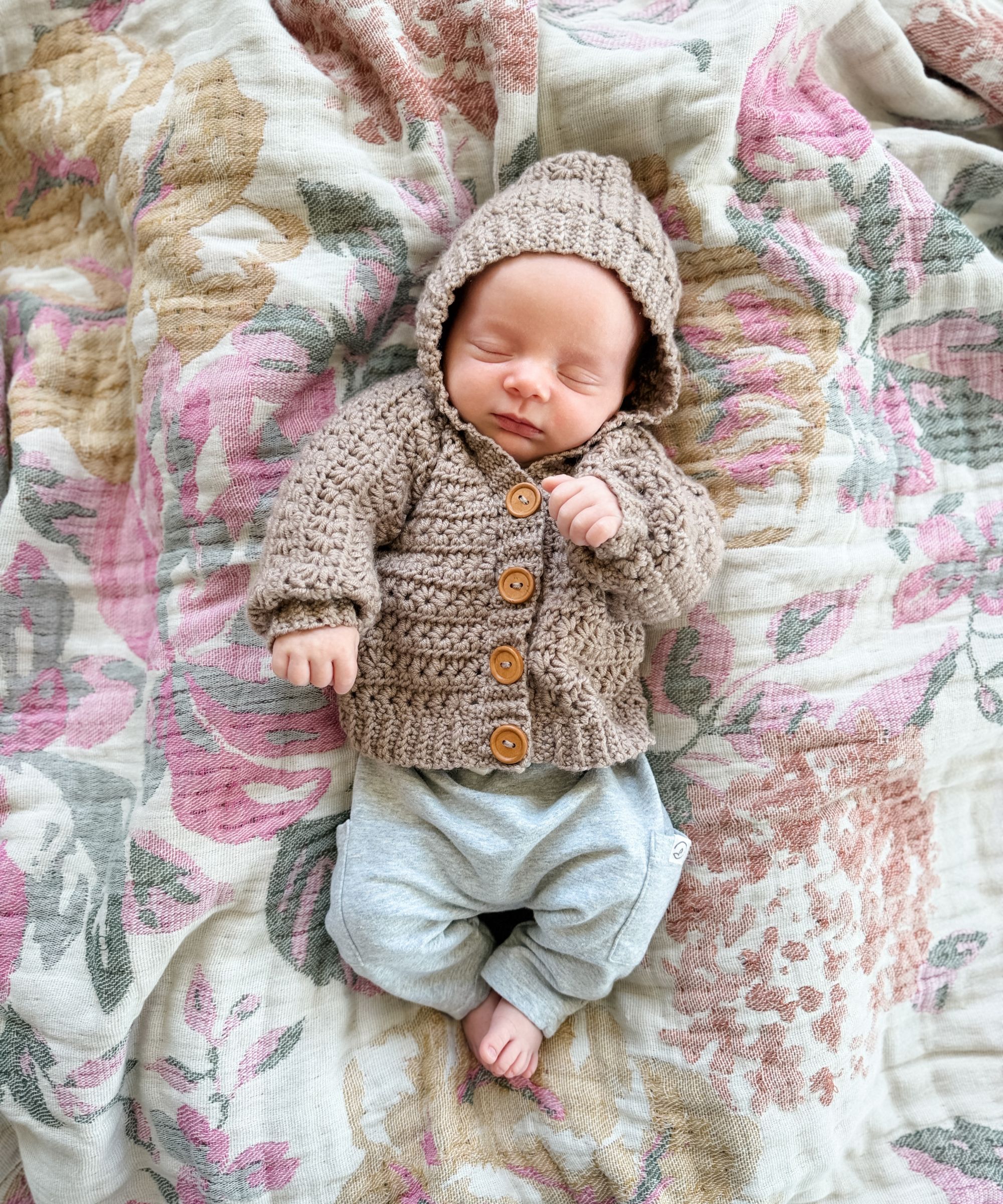 A newborn baby wearing a knitted brown hoodie and gray pants sleeps on a floral-patterned blanket.