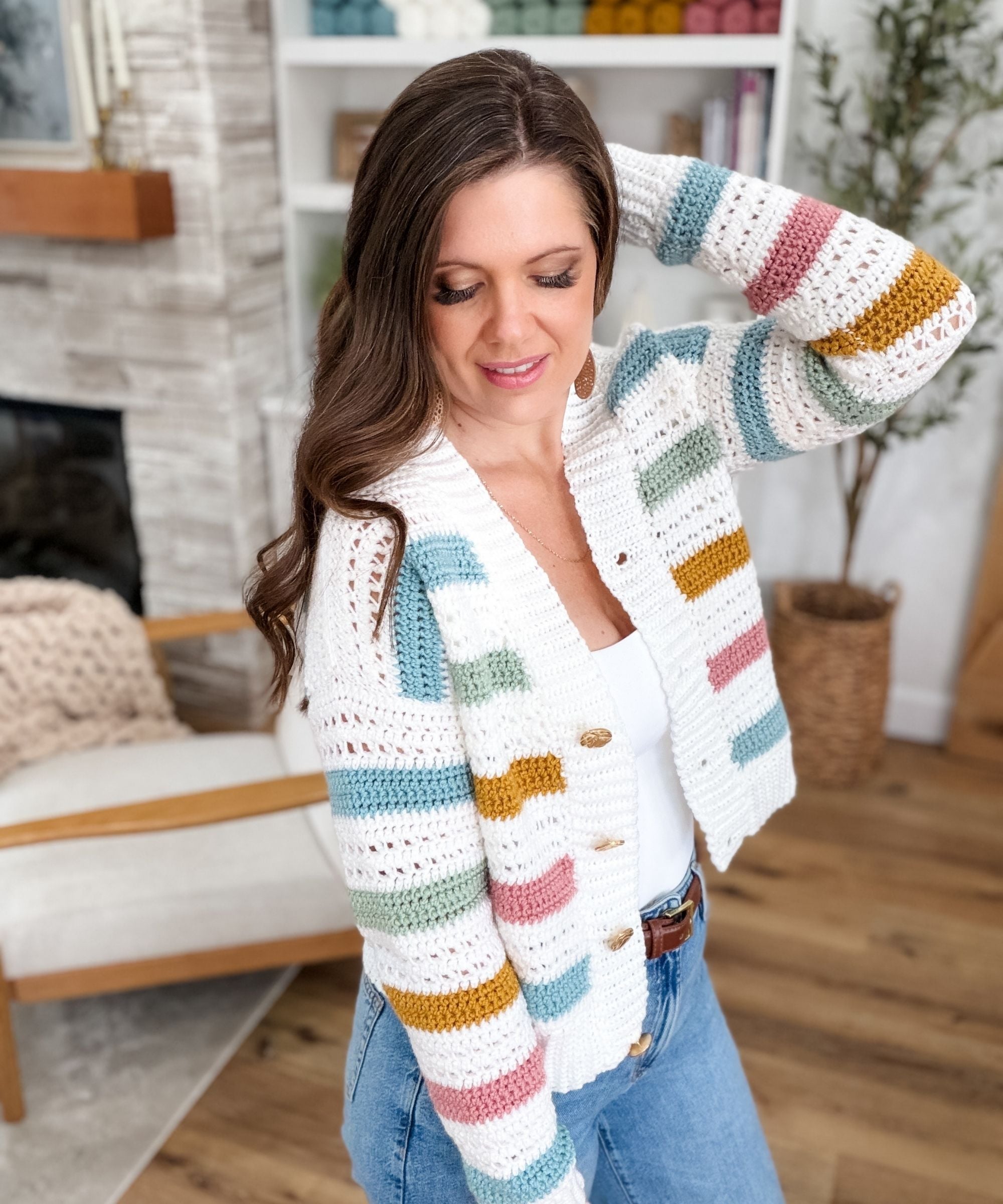 A woman wearing a striped, button-up crochet cardigan stands indoors, touching her hair and looking down, with a cozy chair and plants in the background.