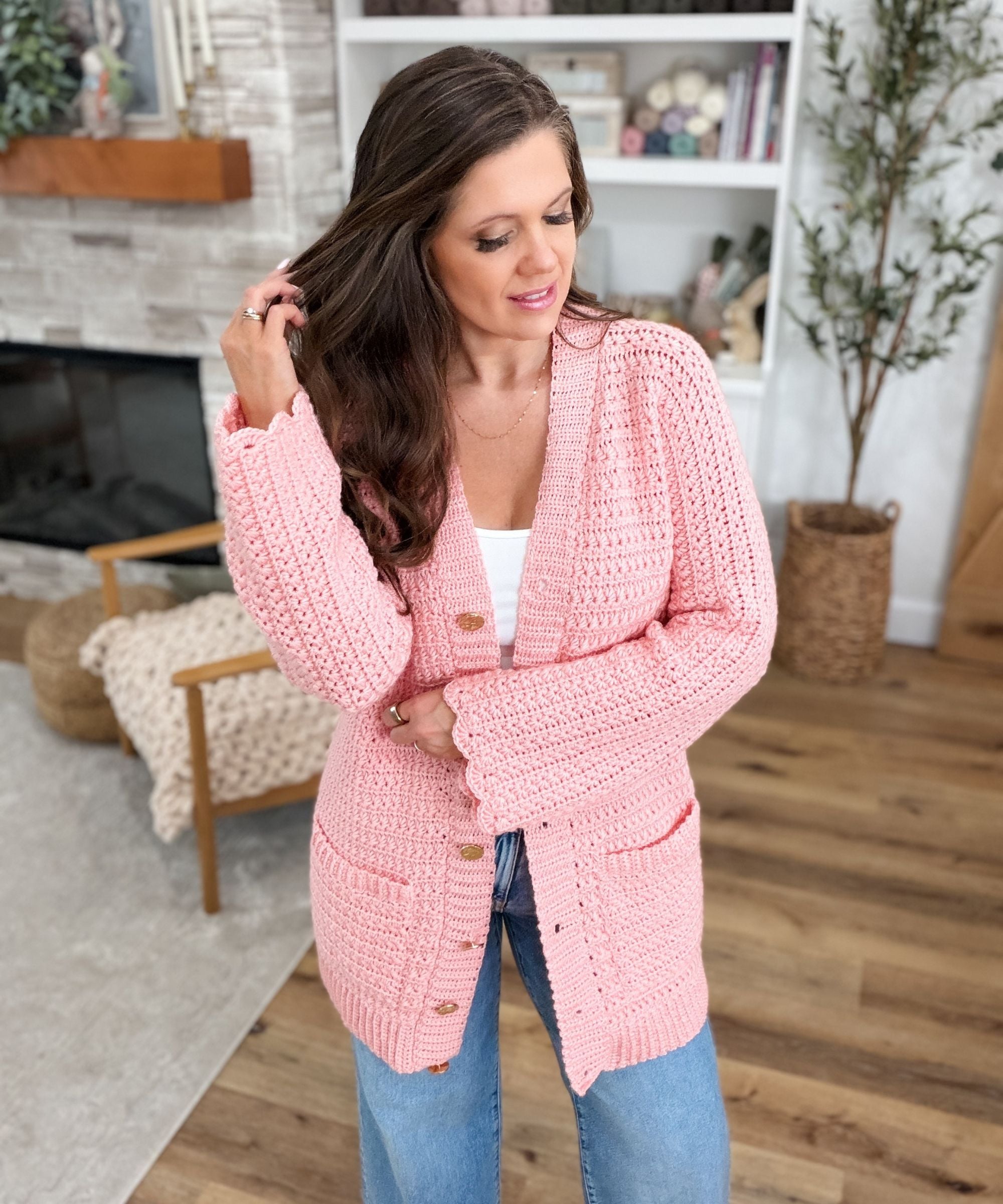 Woman wearing a pink crocheted cardigan over a white top and blue jeans stands indoors, touching her hair, with shelves and a plant in the background.
