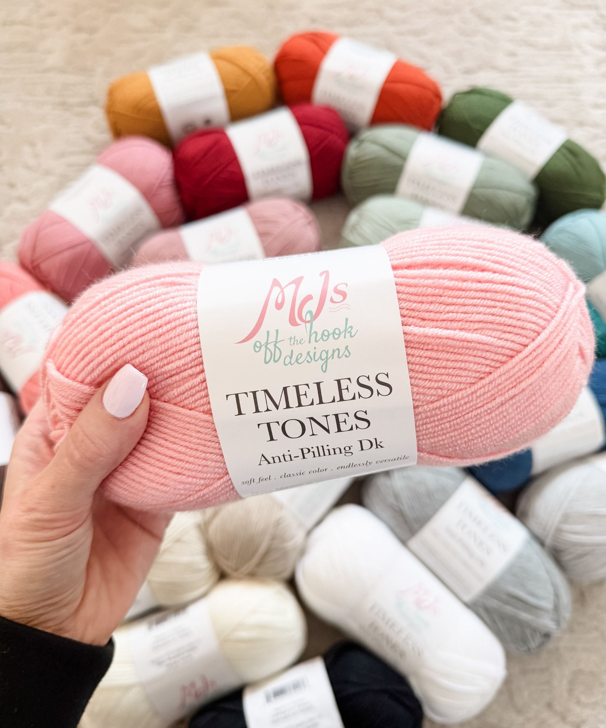 A hand holds a skein of pink "Timeless Tones" anti-pilling DK yarn, with various colored yarn skeins arranged on a carpet in the background.