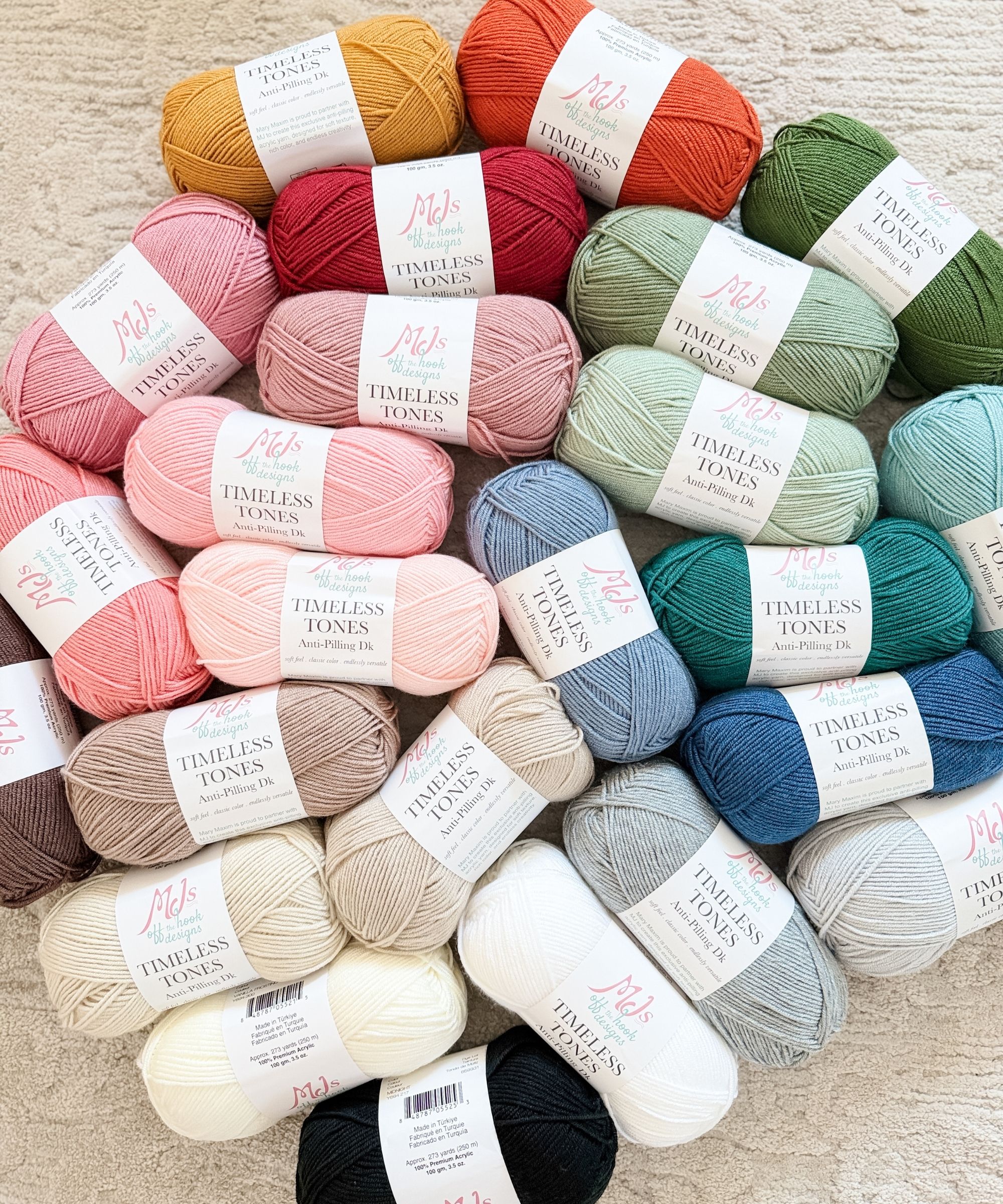 Multiple skeins of Mel's Timeless Tones Yarn Collection, made from soft anti pilling DK yarn in various colors, are arranged on a light-colored carpet.