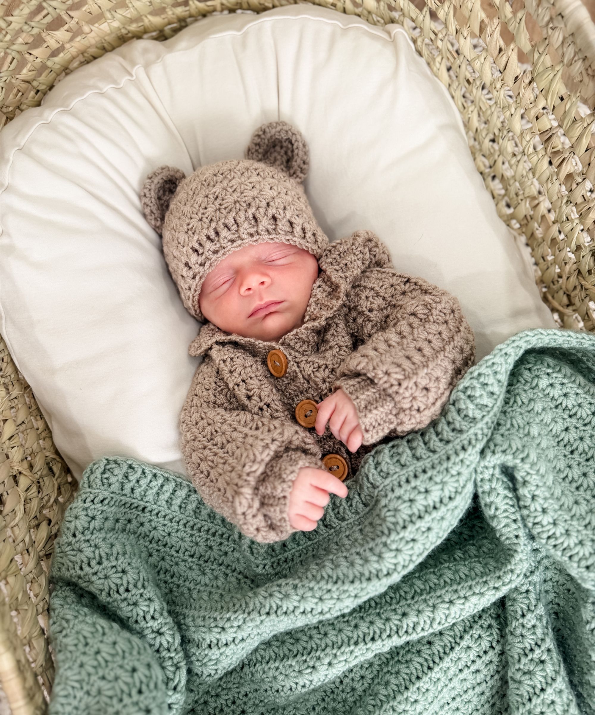 A sleeping baby in a knit bear hat and sweater lies on a white pillow under a green crocheted blanket in a woven basket.