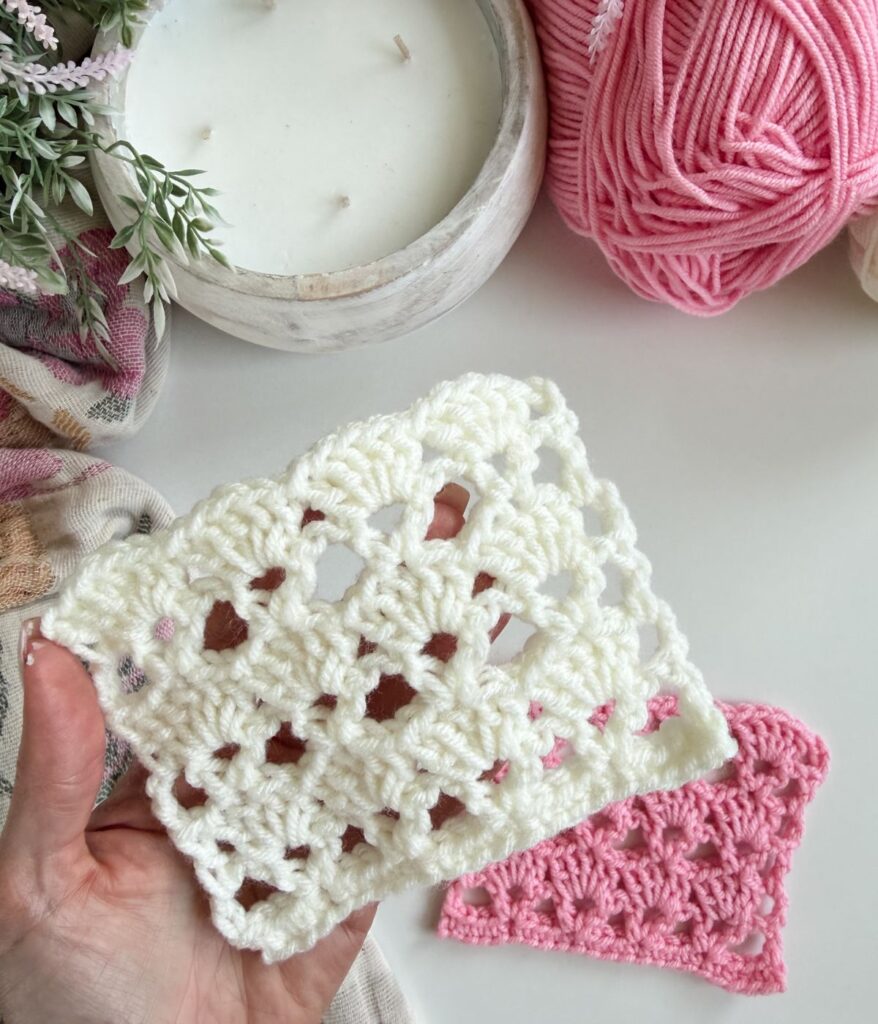 A hand holds a square piece of white crocheted fabric, possibly made with shells crochet; in the background, a pink crocheted square, yarn, and a candle sit on the table.