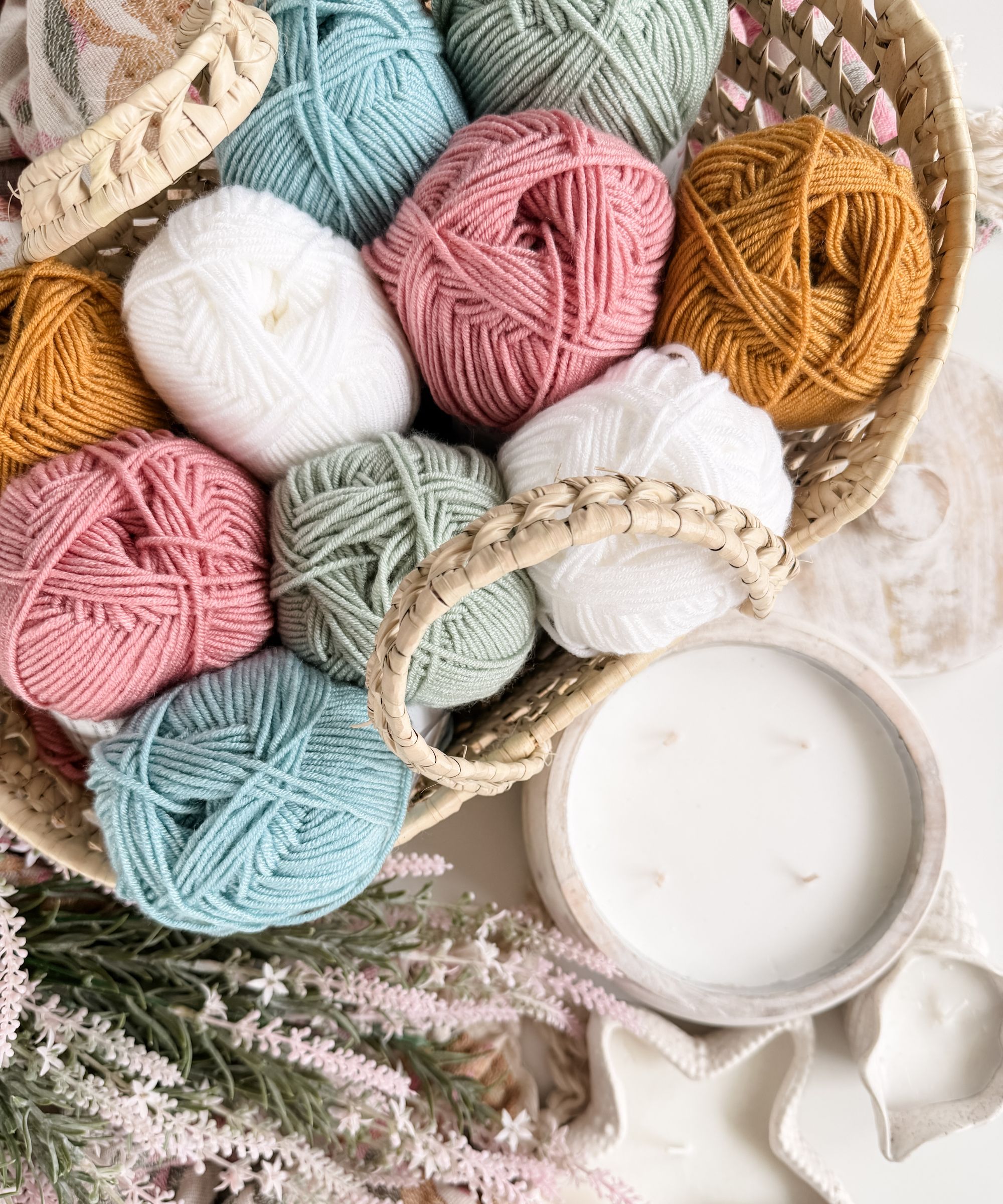 A woven basket filled with colorful yarn balls sits next to a white candle, dried flowers, and seashells on a light surface.