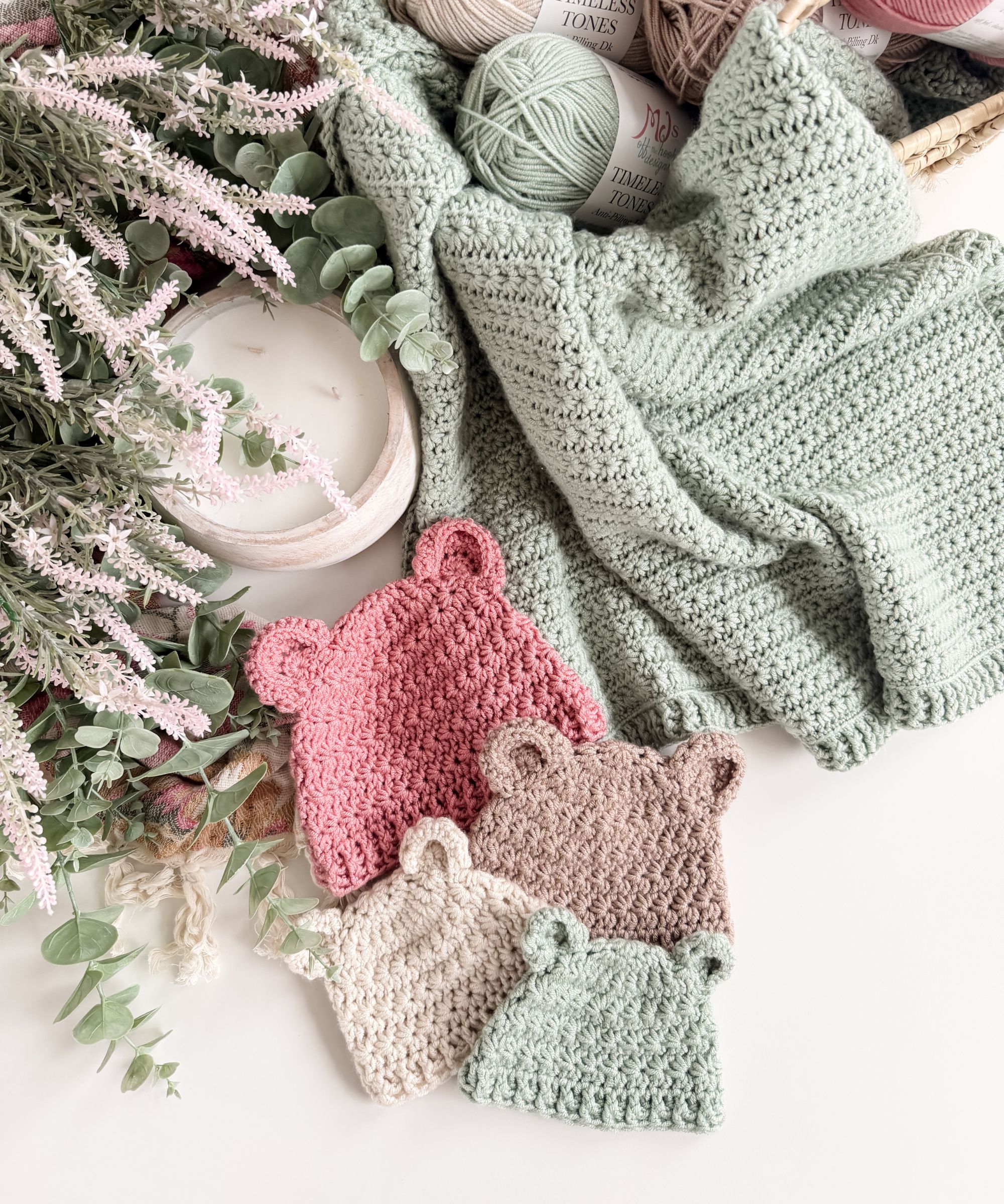 Four crocheted baby hats with bear ears in pink, beige, taupe, and mint are displayed next to a textured mint blanket, yarn, and flowers.