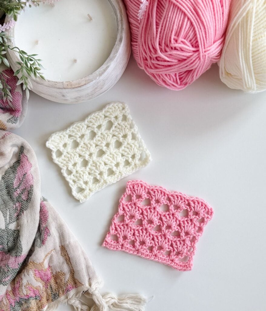 Two crocheted swatches, one white and one pink, featuring v-stitch crochet, are displayed on a white surface near balls of yarn, a candle, and a floral-patterned fabric.
