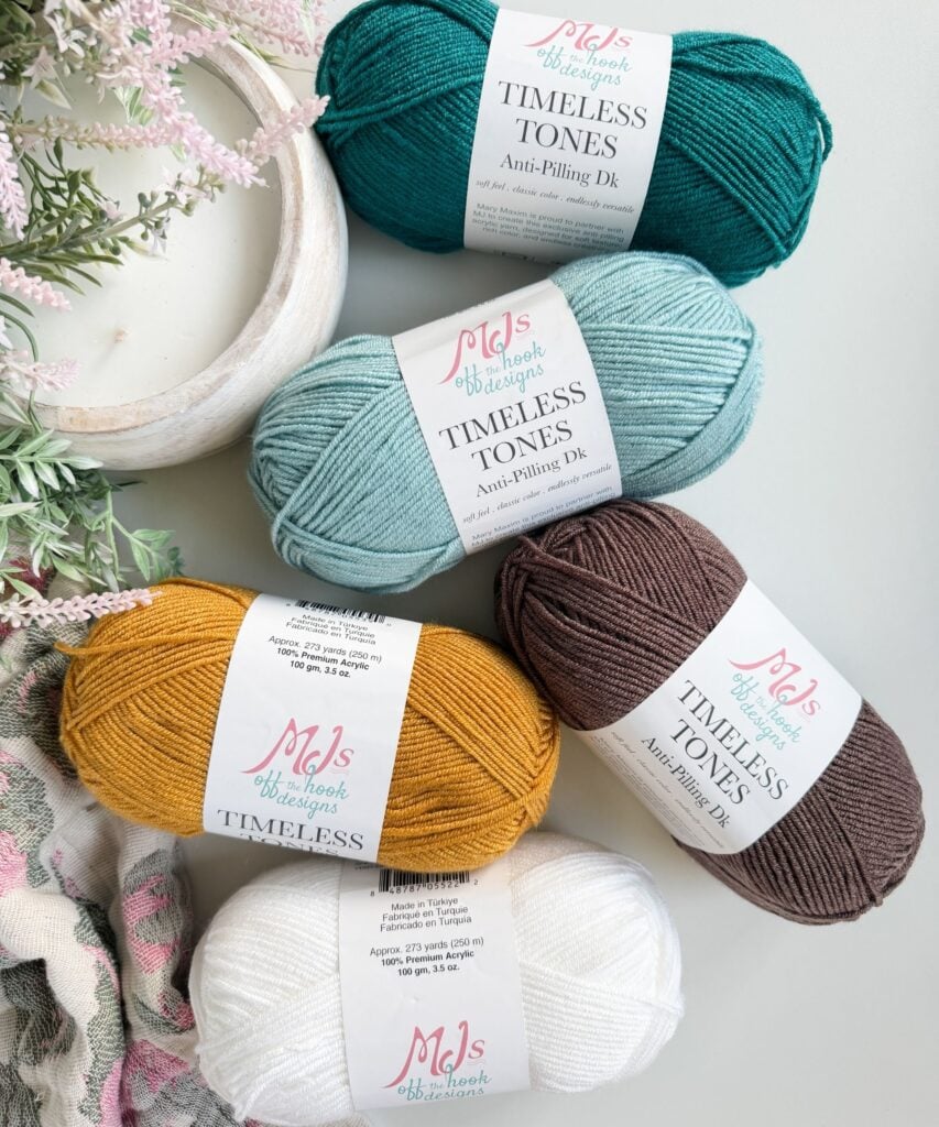 Five skeins of colored yarn (teal, light blue, yellow, brown, and white) labeled "Timeless Tones Anti-Pilling DK" are arranged on a flat surface near a decorative plant.