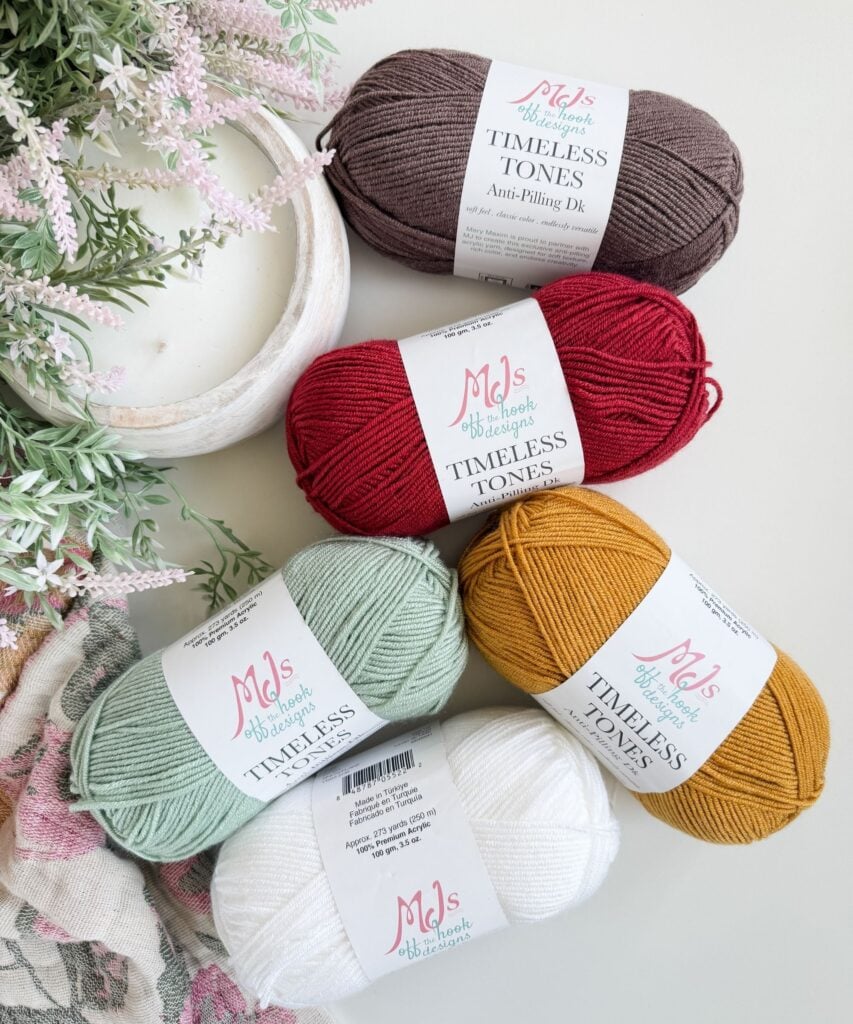 Five skeins of MJ’s Timeless Tones yarn in brown, red, yellow, mint green, and white are arranged on a white surface near flowers and a dish.
