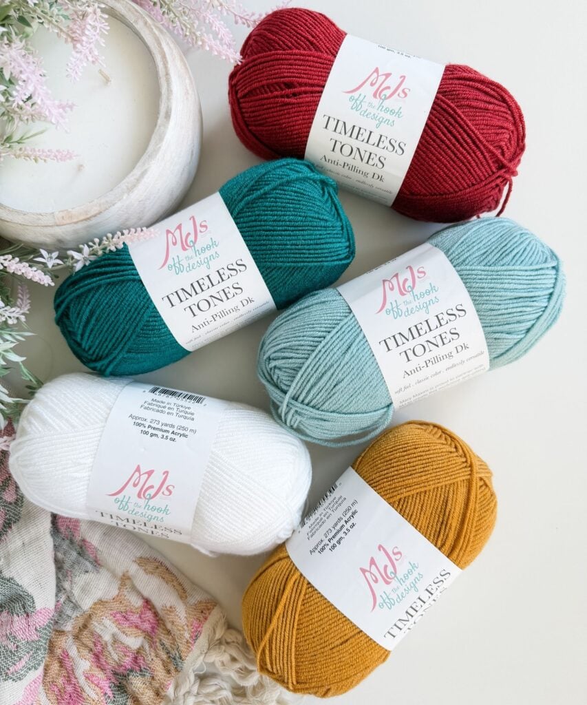 Five skeins of MJ’s Timeless Tones yarn in red, teal, light blue, white, and mustard yellow are arranged on a white surface next to a floral textile and a plant.