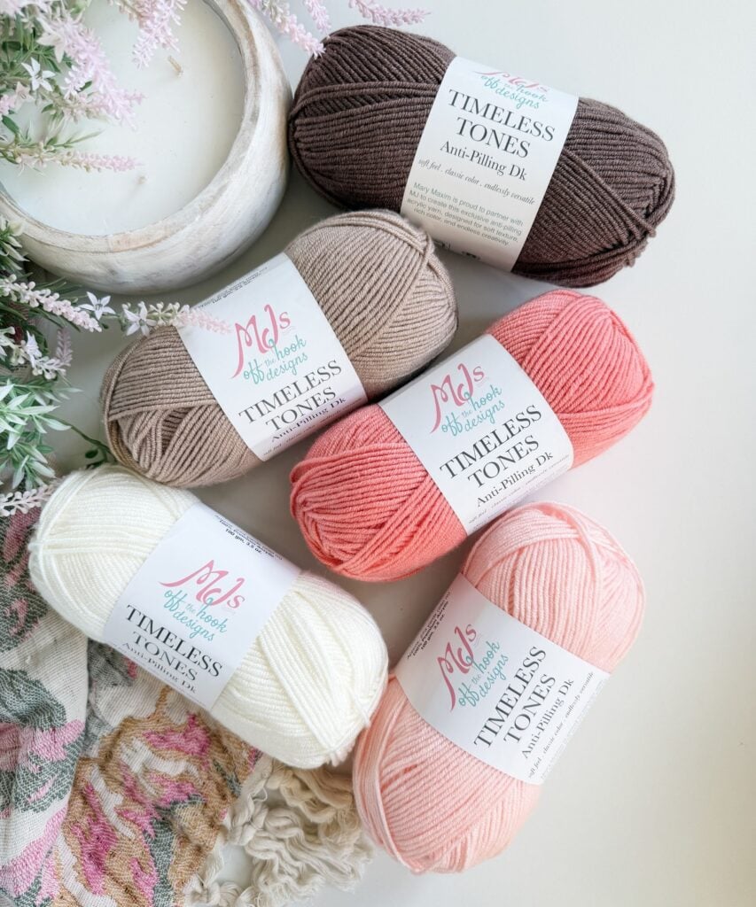 Five skeins of yarn in brown, beige, pink, peach, and white, labeled “Timeless Tones Anti-Pilling DK,” are arranged on a white surface next to a floral fabric and plant.