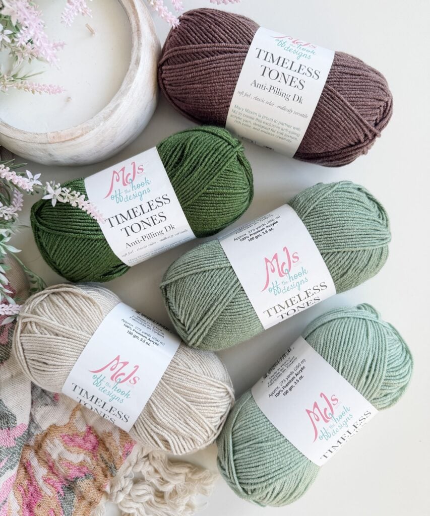 Five skeins of "Timeless Tones" yarn in various earth tones are arranged on a white surface with some decorative flowers visible in the background.