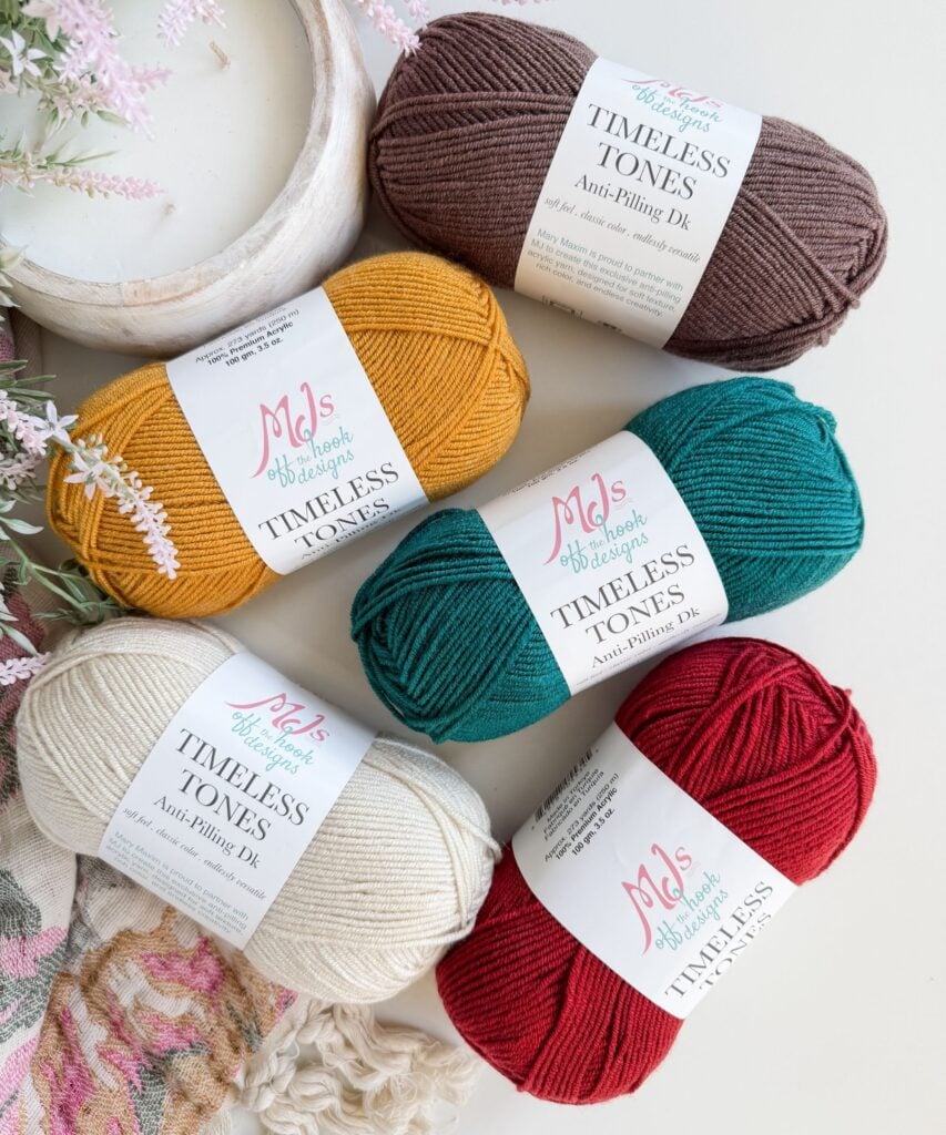 Five skeins of anti-pilling yarn in brown, mustard yellow, teal, cream, and red are arranged on a white surface with pink florals and a patterned fabric nearby.