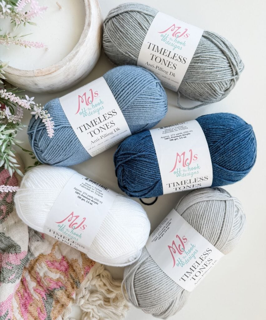 Four skeins of anti-pilling yarn in shades of blue, grey, and white are arranged next to a floral fabric and decorative flowers.