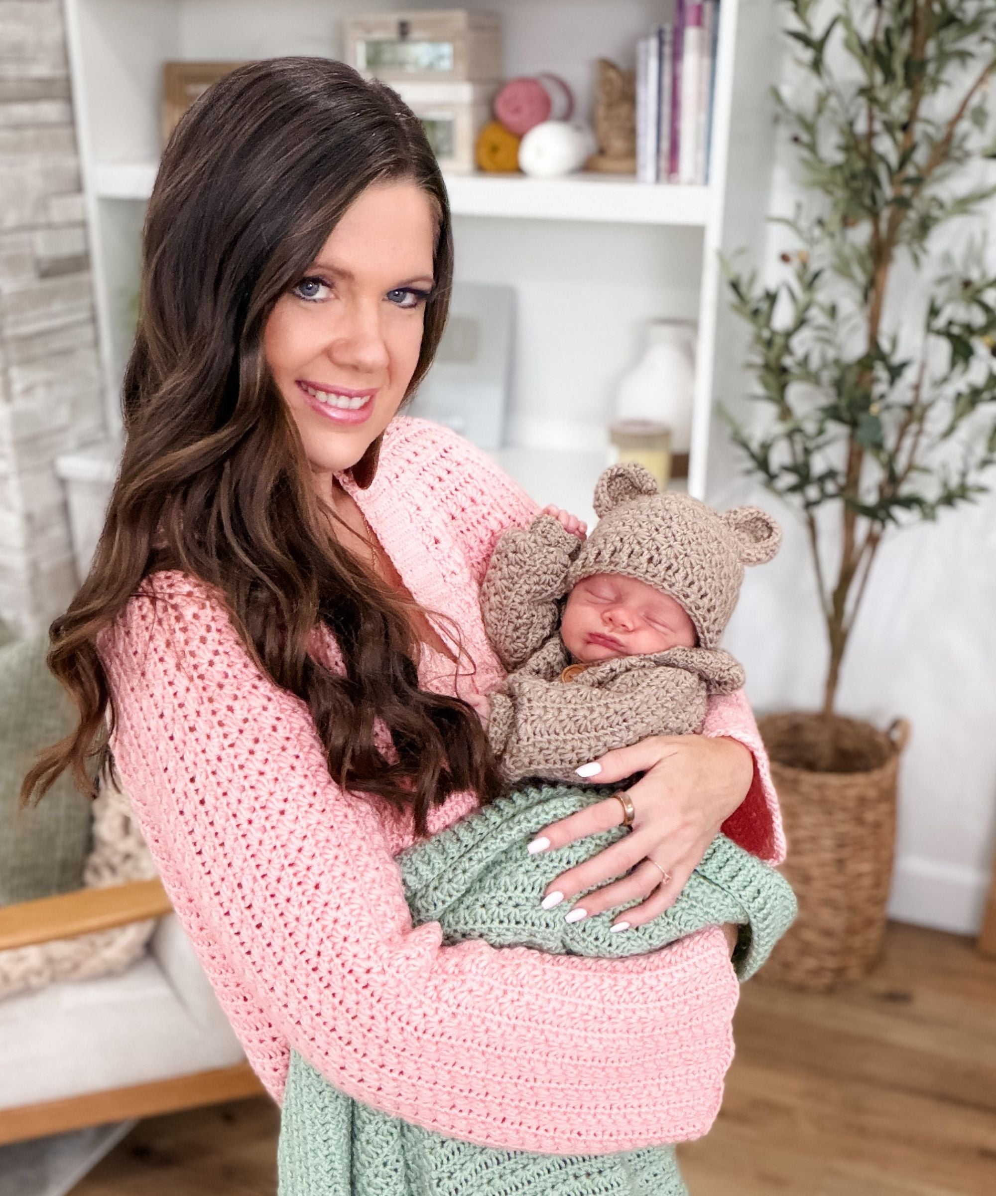 Woman in a pink sweater holds a newborn baby dressed in a brown knit bear outfit and green blanket, standing in a cozy room with shelves and plants in the background.