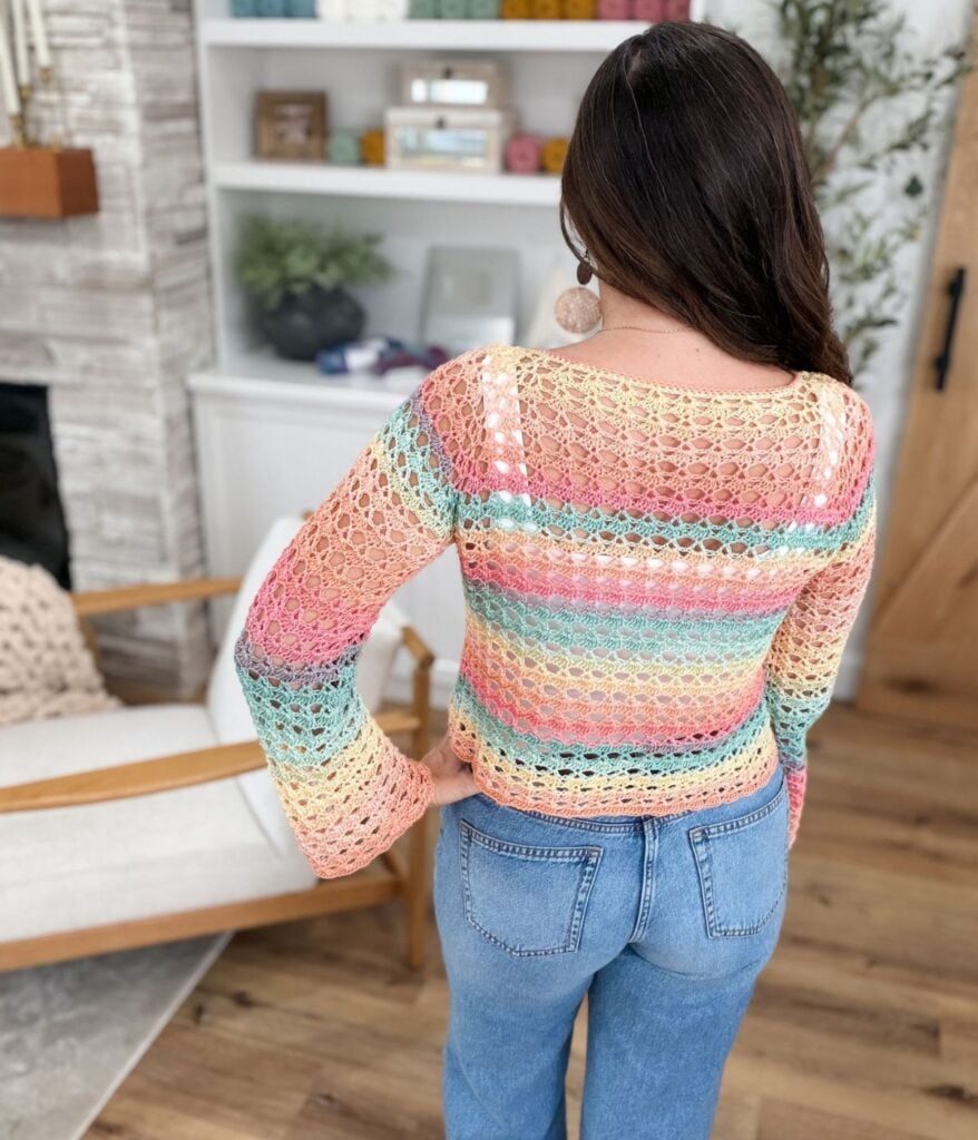 A woman with long brown hair wears a colorful crocheted sweater, made with a shell stitch crochet pattern, and blue jeans, standing with her back to the camera in a living room with shelves and a chair.