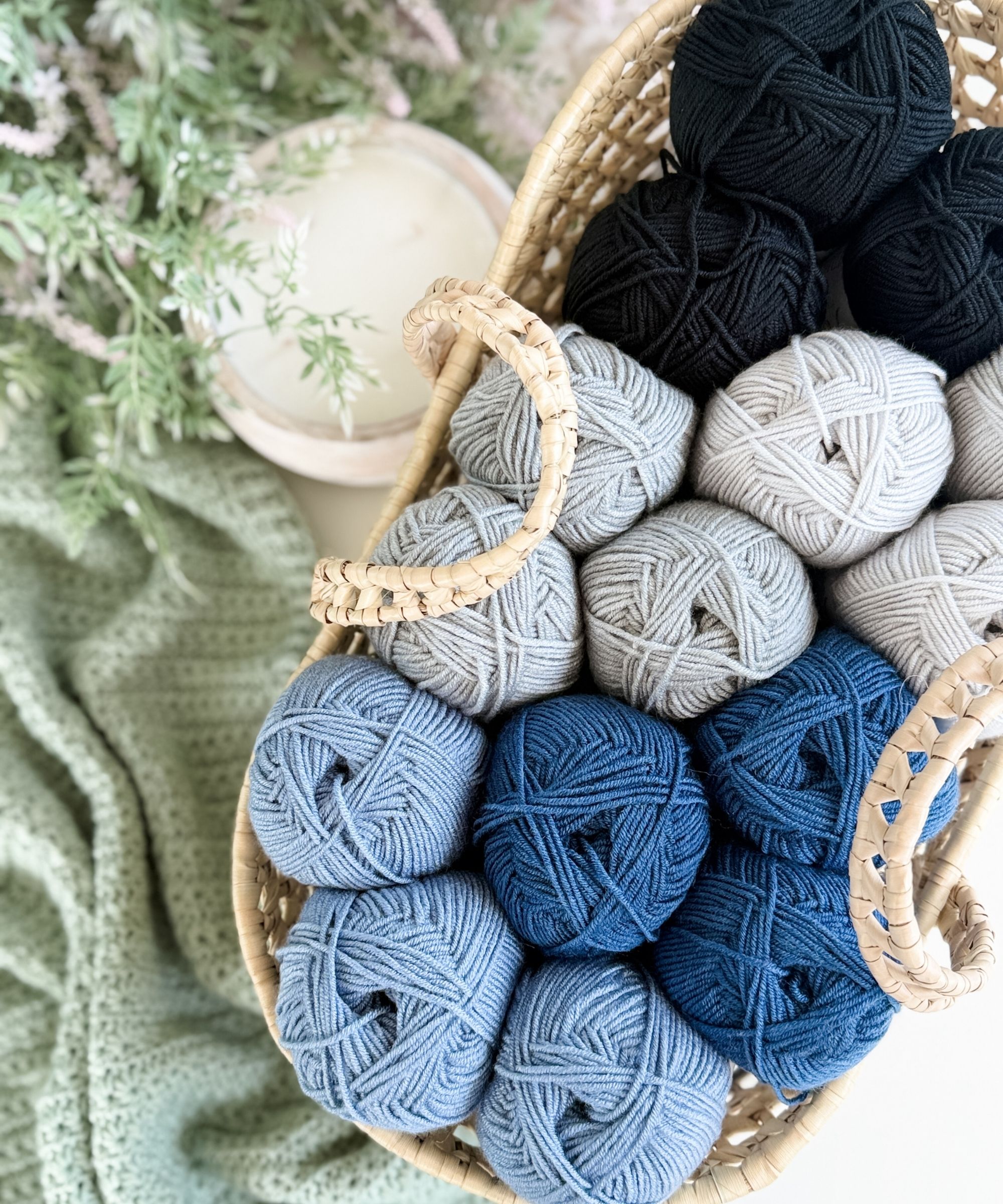 A woven basket holds balls of yarn in shades of black, gray, and blue, next to a crocheted green fabric and a leafy plant.