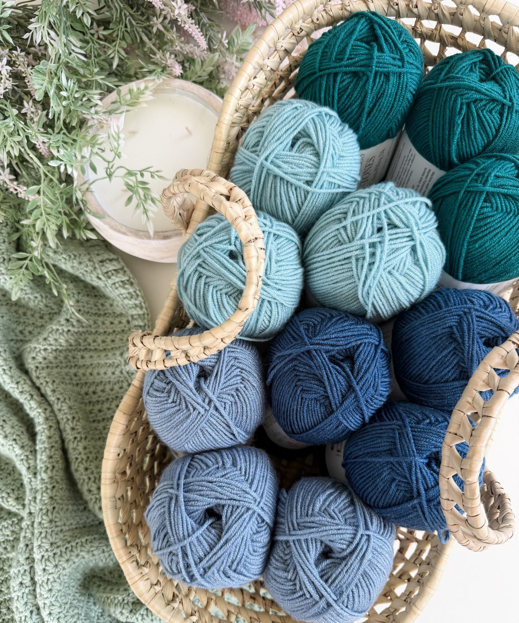A woven basket holds skeins of yarn in shades of green and blue. A knitted fabric, a plant, and a candle are nearby on a white surface.