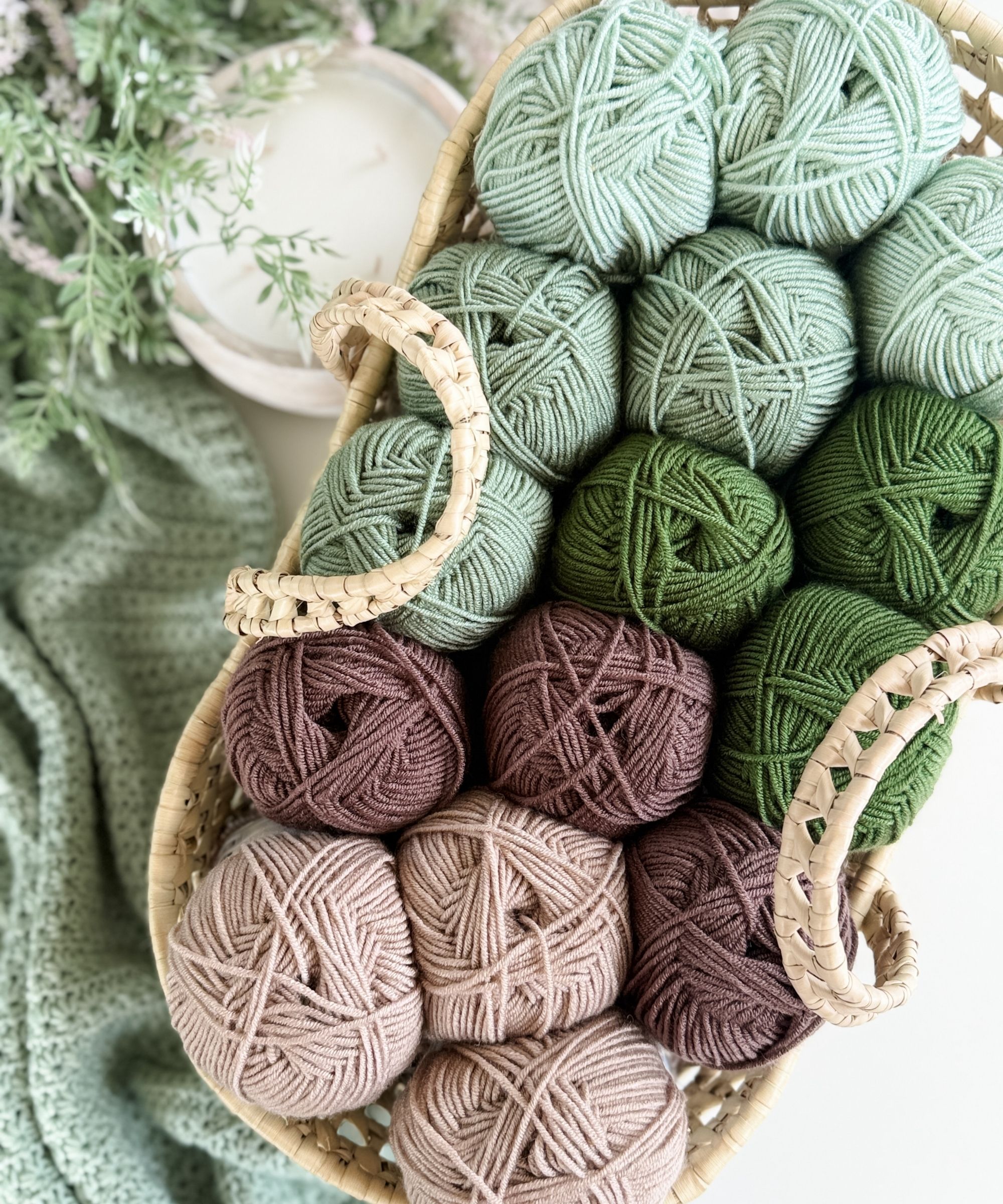 A woven basket filled with balls of yarn in shades of green and brown sits beside a green crocheted fabric and a white candle.