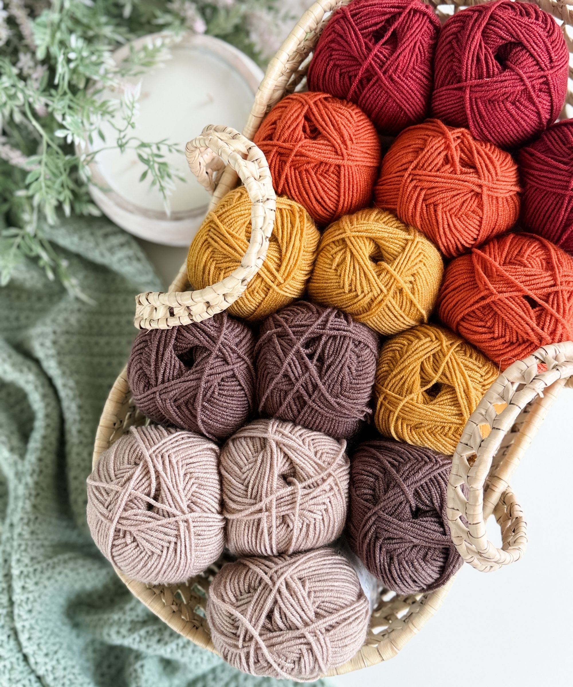 A woven basket holds neatly arranged yarn balls in shades of red, orange, yellow, brown, and beige, next to a green knitted blanket and a plant.