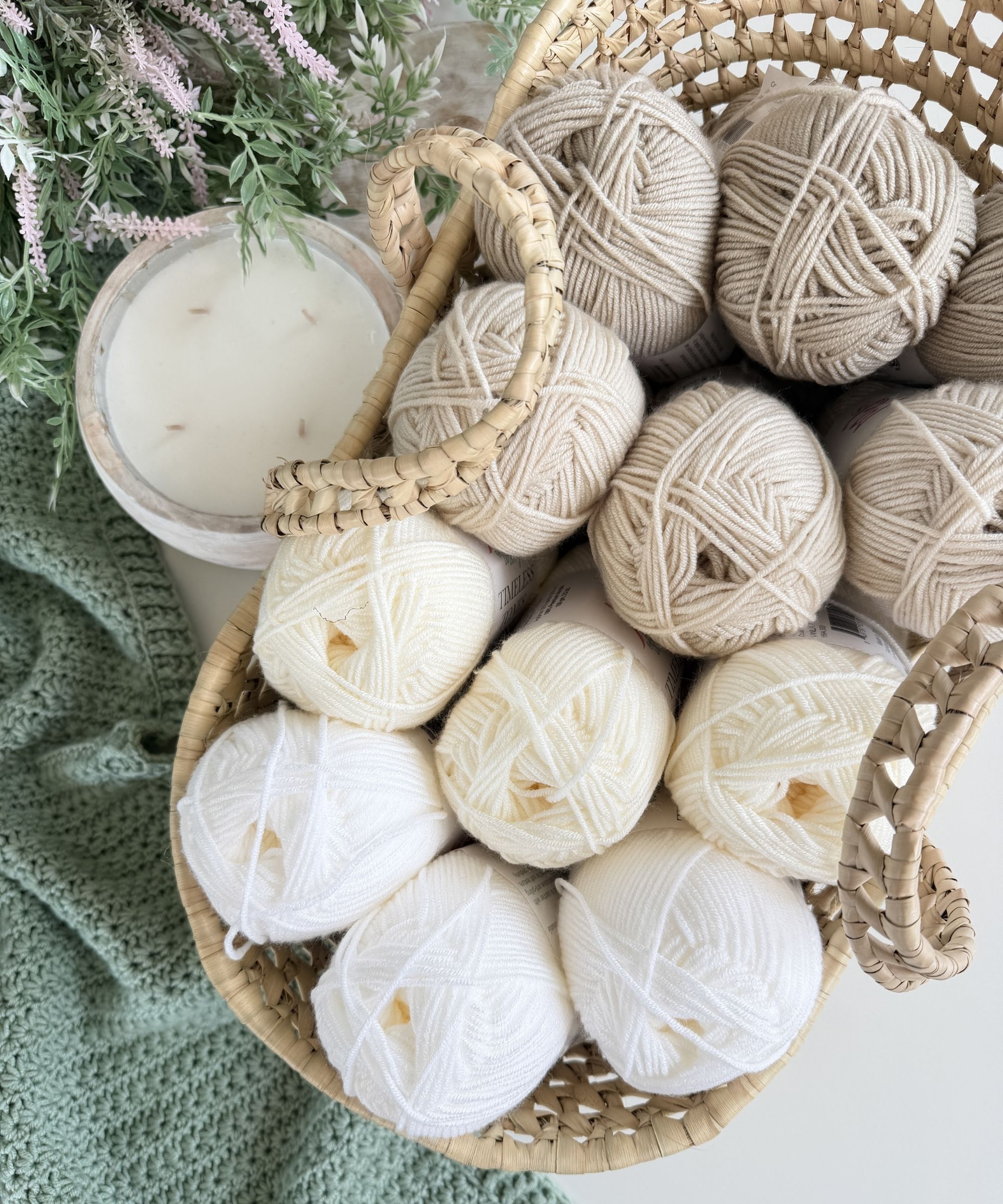 A woven basket filled with white and beige yarn balls sits next to a green crochet piece, a white candle, and some leafy greenery.