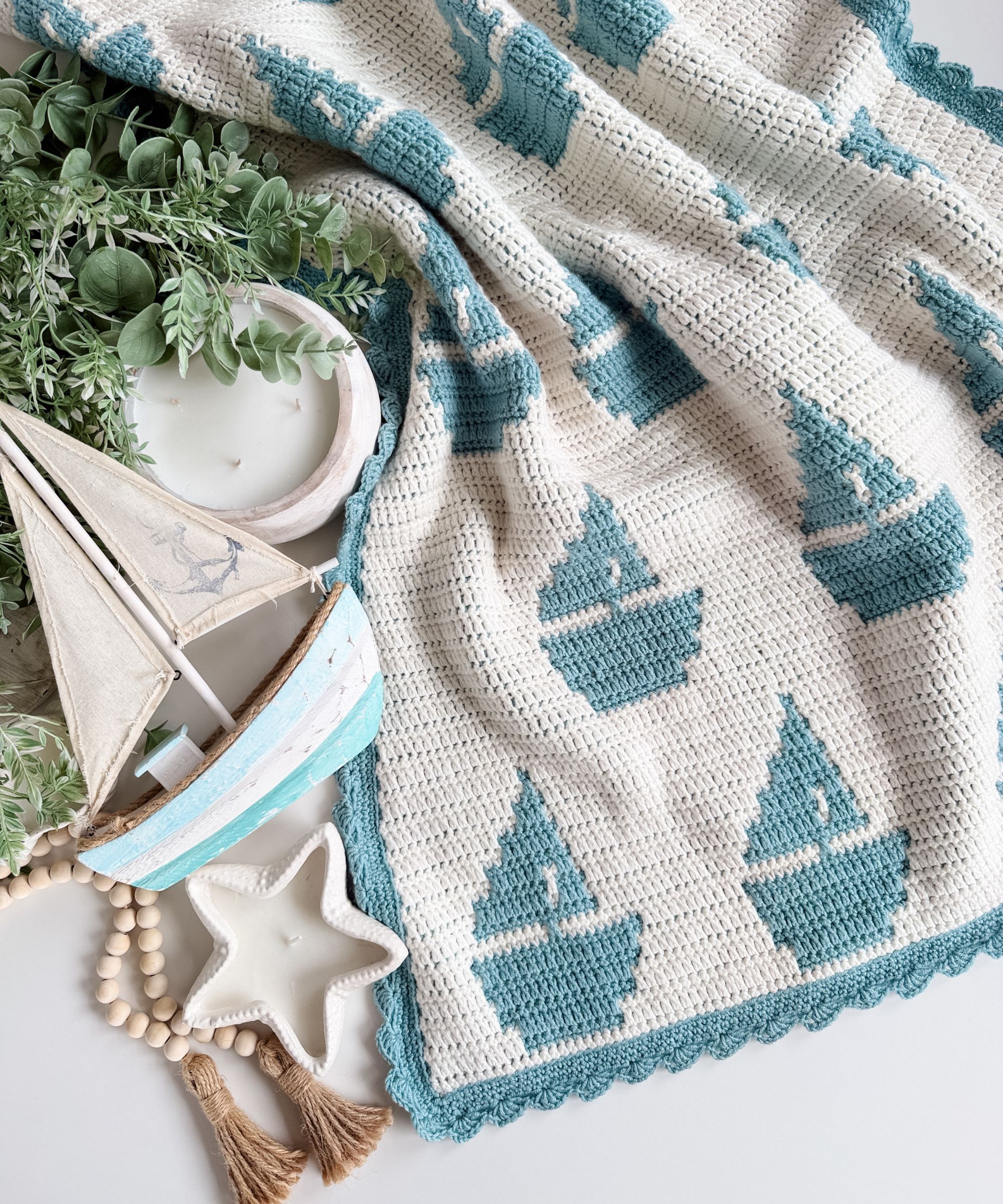 A crocheted blanket with alternating teal sailboat and sea creature patterns, displayed with a wooden sailboat, greenery, beads, star-shaped dish, and candle.