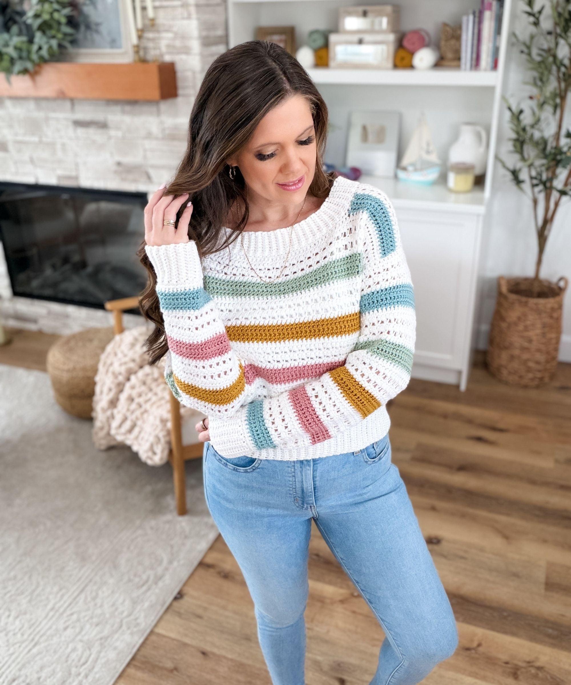 Woman with long brown hair wearing a striped sweater and blue jeans stands indoors, touching her hair, in a cozy living room with wooden floors and shelves in the background.