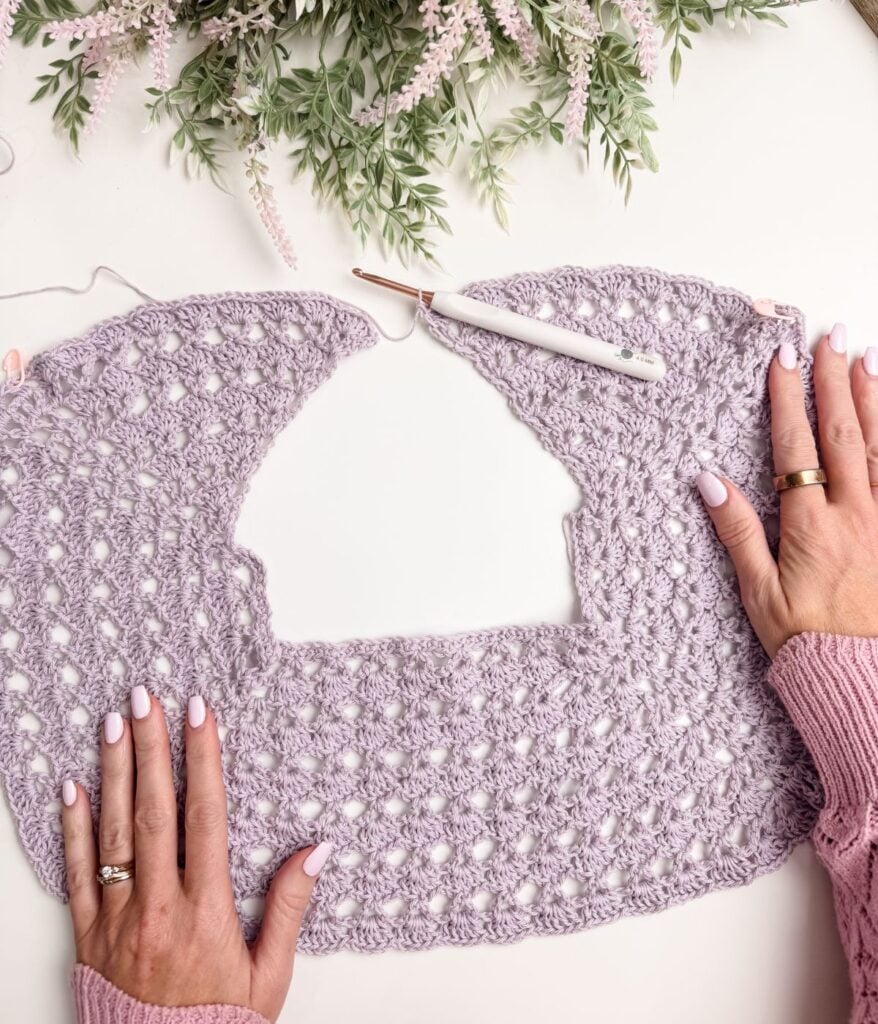 Hands hold a partially completed lavender crochet top pattern with a white crochet hook, on a white table decorated with green and pink foliage.